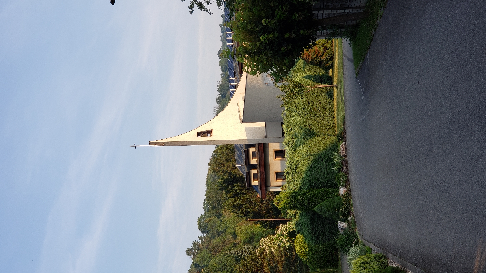 A modern church with a distinctive triangular bell tower is situated on a gentle slope, partially obscured by lush green trees and manicured hedges. The church's facade is a clean white, contrasting with the darker roofs and windows of the adjacent buildings. Solar panels are visible on the rooftops, suggesting a commitment to renewable energy. A paved road curves in the foreground, leading towards the church. The sky is a clear, pale blue with faint wisps of clouds. The overall scene suggests a peaceful, well-maintained suburban environment. The church is the Roman Catholic Church of St. Nicholas, located in the Sekčov district of Prešov, Slovakia.