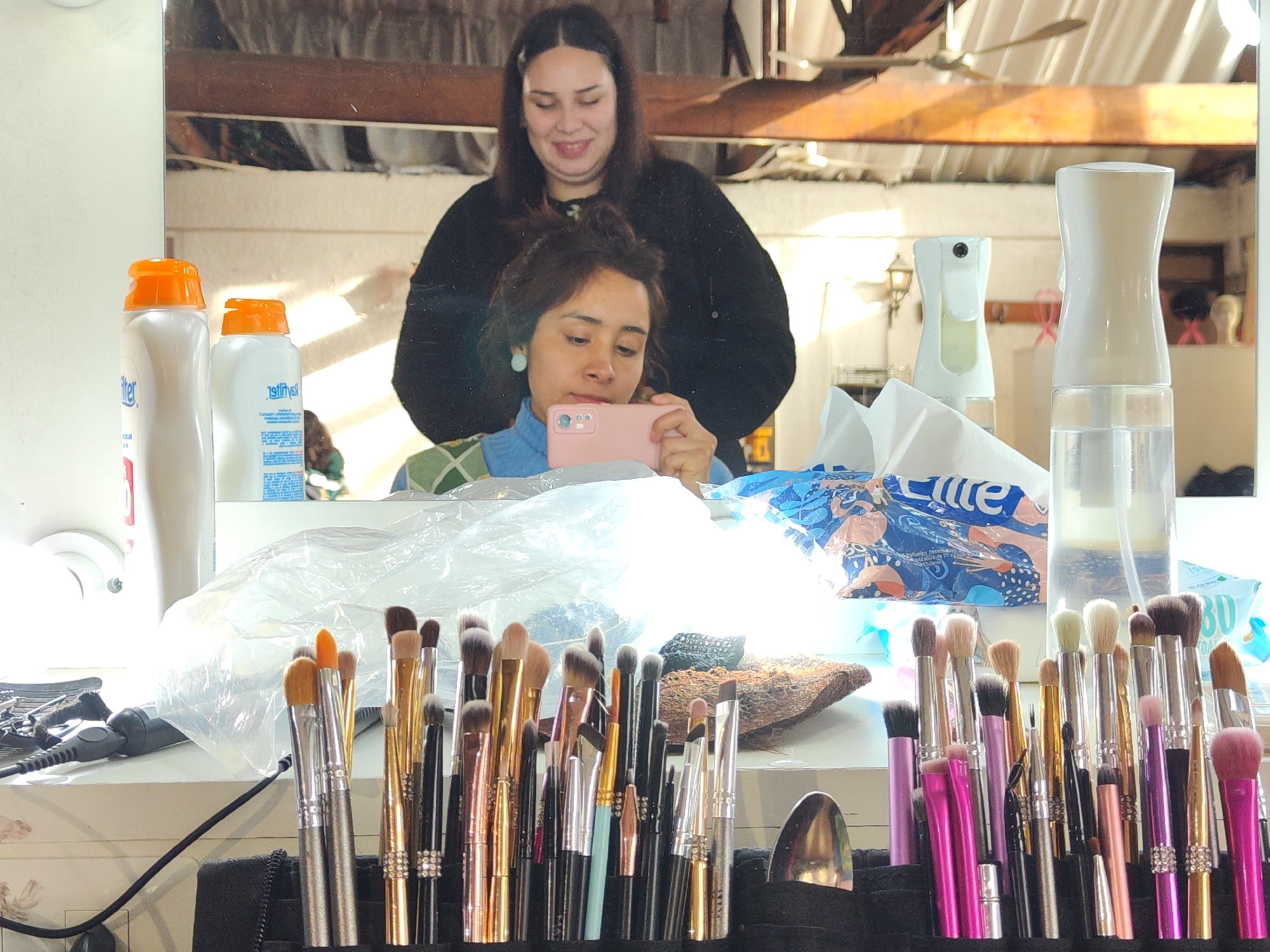 The image depicts an indoor scene, likely a vanity area or makeup studio, with two individuals reflected in a mirror. The primary focus is on a collection of makeup brushes arranged in a black holder in the foreground. Behind the brushes, various cosmetic products are visible, including two spray bottles with orange caps, a larger white spray bottle, and a package of what appears to be tissues or wipes.
In the reflection, a young woman is holding a pink smartphone and looking at it. She is wearing a blue turtleneck sweater with a checkered overlay. Behind her, another woman is reflected, wearing a black top and smiling. The background of the reflection shows a ceiling with exposed beams and a fan, suggesting a room with an industrial or rustic aesthetic. The lighting appears to be natural, possibly from a window, casting bright highlights on the objects.
While the image is rich in detail about the makeup setup and the individuals, there is no information provided within the image itself to indicate its specific location in Santiago, Chile.