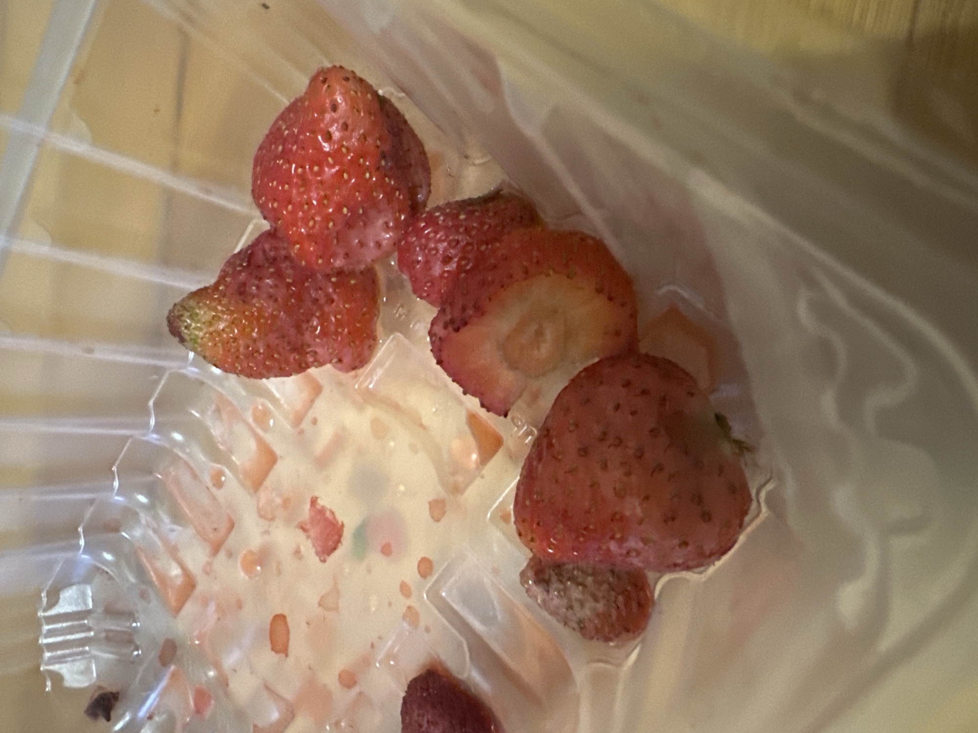 The image shows a partially eaten container of strawberries in a plastic clamshell. The container is clear plastic with individual compartments. Several strawberries, some whole and some cut, are scattered within the container. There are also remnants of strawberry juice and pulp visible. The background appears to be a wooden surface. There is no indication of the city of Saransk, Russia in the image.
