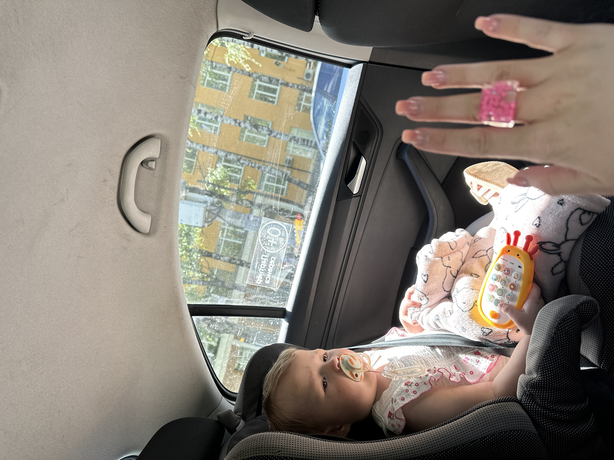 The image captures a view inside a car, focused on a baby in a car seat, with an urban scene visible through the window.
In the foreground, a baby lies in a grey car seat, looking upwards with a pacifier in its mouth. The baby is dressed in a light-colored outfit, possibly white with pink accents, and is partially covered by a pink patterned blanket. Its right hand holds a bright yellow toy phone, shaped like a giraffe, featuring numerous colorful buttons and a small screen displaying "0+" and "12:00".
Above the baby, an adult's hand is visible, blurry and partially cropped, wearing a prominent chunky pink ring. The hand has light pink painted nails.
The car's interior details include a light grey headliner with a white grab handle on the left, and a dark grey door panel with a speaker grill on the right. Through the car window, a multi-story building with a yellow-ochre facade and numerous windows is clearly visible. Green foliage from trees can also be seen, both directly and reflected in the building's windows. The bright sunlight suggests it is daytime.
A sticker on the car window, appearing mirrored and upside down, displays "0+" and partially legible Cyrillic text, possibly including "СОДЕРЖИТ" (contains) and "безопасность" (safety). The scene depicts a calm baby on a car journey within an urban setting, consistent with the provided location context of Nizhny Novgorod, Russia.