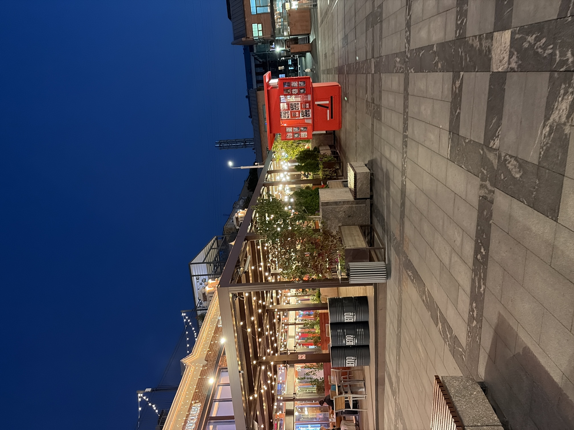 The image depicts an outdoor scene at dusk or night, likely in an urban area. The sky is a deep, clear blue. In the foreground, a paved walkway with a decorative pattern of darker stone is visible. To the left, a portion of a concrete and wooden bench is seen.
The main subject is a restaurant or cafe with a covered outdoor seating area. String lights illuminate the space, casting a warm glow on the tables and chairs. Several large, black barrels with white lettering are stacked near the entrance to the outdoor seating area. To the right of the restaurant, a bright red, kiosk-like structure stands, adorned with photographs. Beyond this, there are more buildings, some with windows lit from within, and streetlights. A single person is visible walking in the distance.
The architecture suggests a modern or renovated urban space, possibly a public square or a pedestrian zone associated with entertainment or dining venues. The overall atmosphere is calm and inviting, characteristic of a public gathering place in the evening.
While the exact location in Moscow cannot be confirmed from the visual cues alone, the presence of Cyrillic lettering on the barrels and the architectural style are consistent with urban settings in Russia.