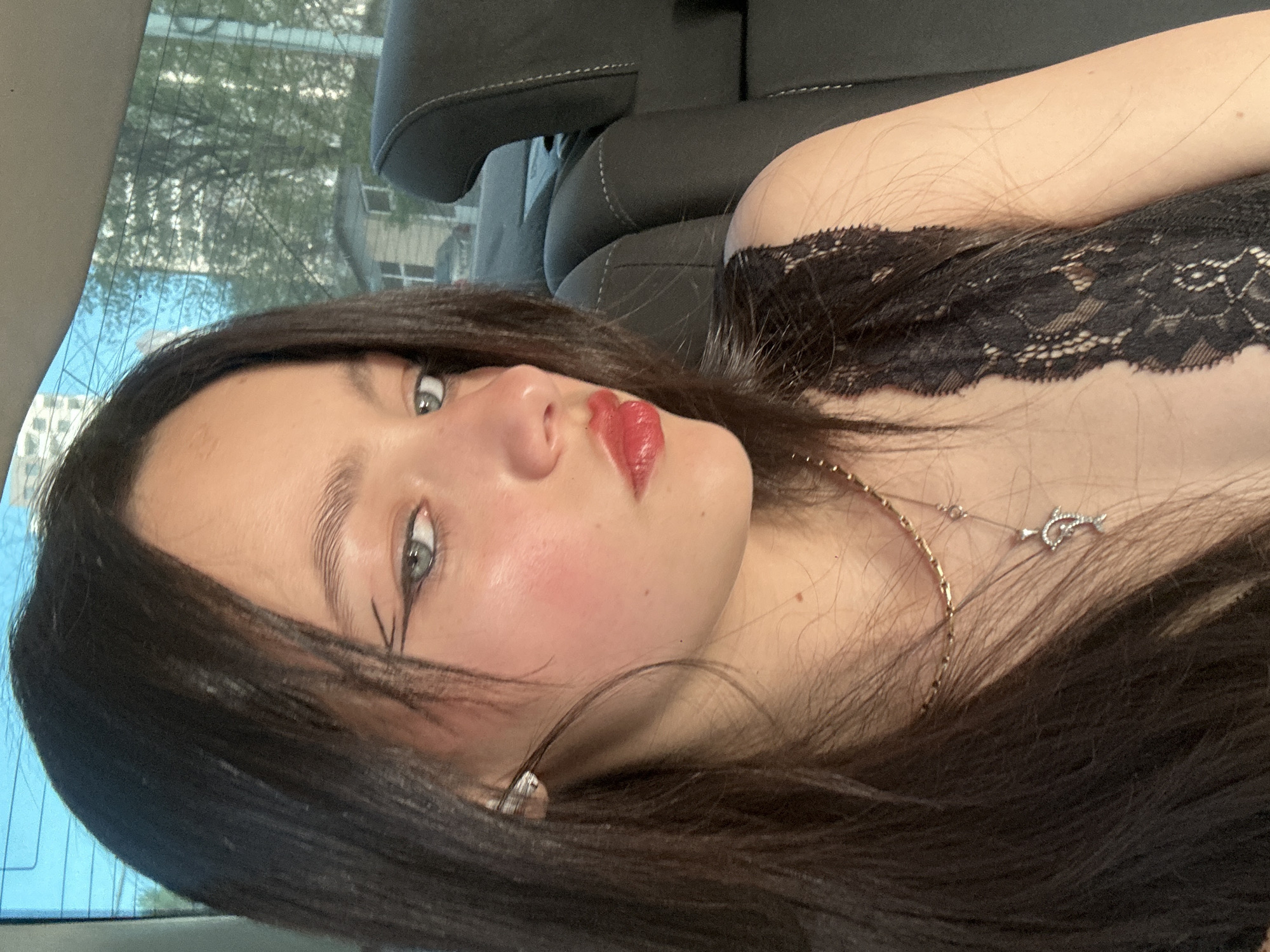 A young woman is lying in the passenger seat of a car, looking at the camera. She has dark hair, fair skin, and is wearing red lipstick and makeup. She is wearing a black lace top and a gold necklace with a silver dolphin pendant. The interior of the car is visible, with black leather seats. A cityscape is visible through the car window. It is not possible to verify the location within the city of Yerevan based on the image.