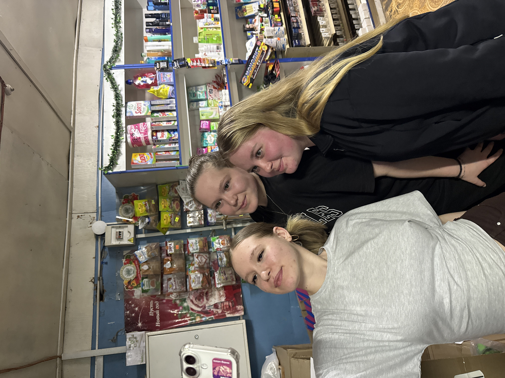 Three girls are posing for a selfie in what appears to be a small convenience store. The store has shelves stocked with various goods, including food items and household products. A Christmas garland hangs on one of the shelves. A poster featuring Santa Claus and text in Russian, potentially related to the New Year, is visible on the wall. The walls are painted in light blue. The camera is a smartphone with a clear case. The location is Rusalevka, Russia.