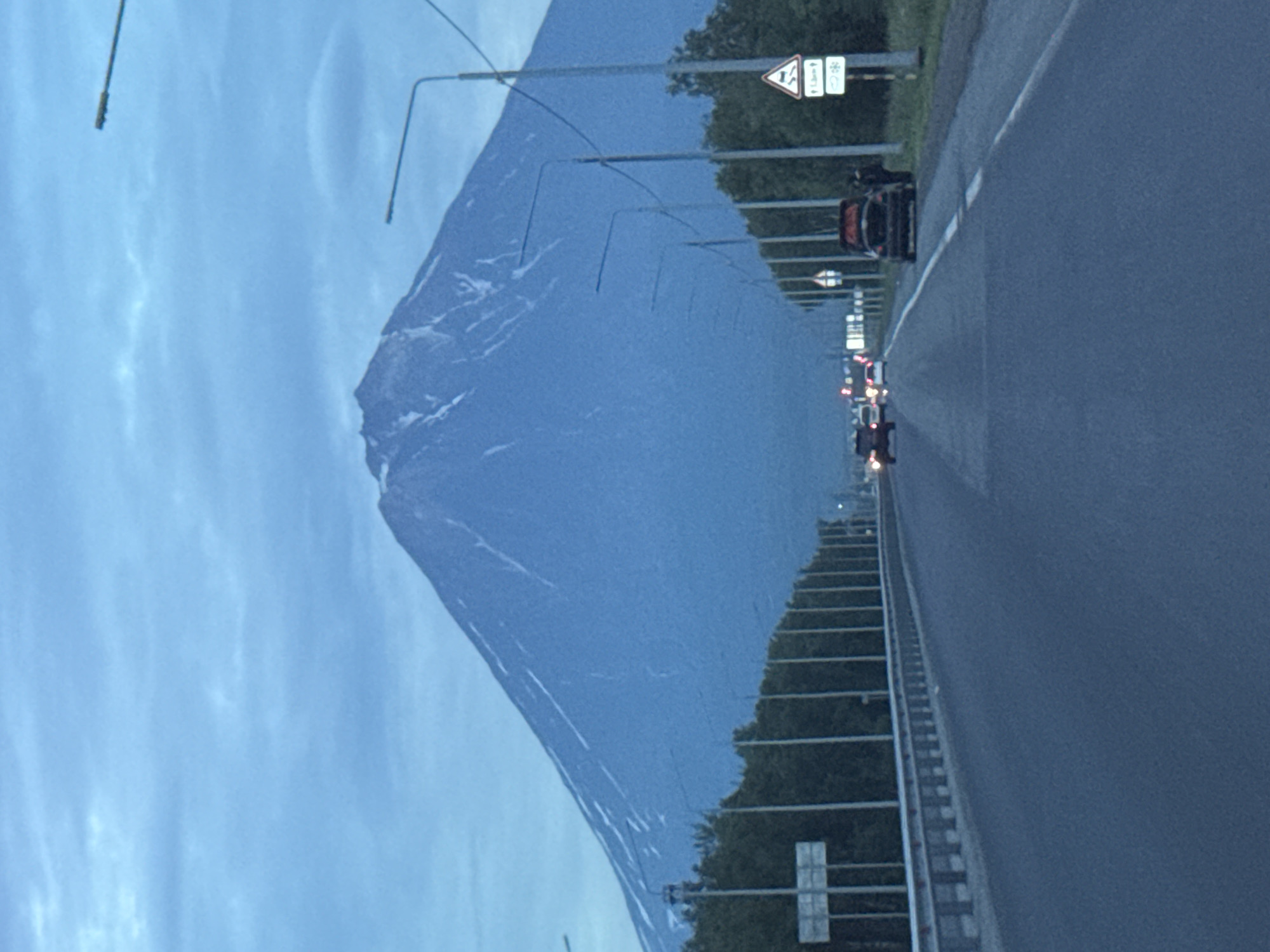In Yelizovo, Russia, on a cloudy day, a vast blue mountain with patches of snow dominates the background. In the foreground, a two-lane road stretches towards the mountain, lined with trees and streetlights. Several cars are visible on the road, with one black car stopped on the shoulder, its trunk open, and a person standing beside it. A traffic sign indicating a sharp turn and a weather warning is posted nearby. The overall scene suggests a scenic drive near a volcanic region, possibly at dawn or dusk due to the low light.