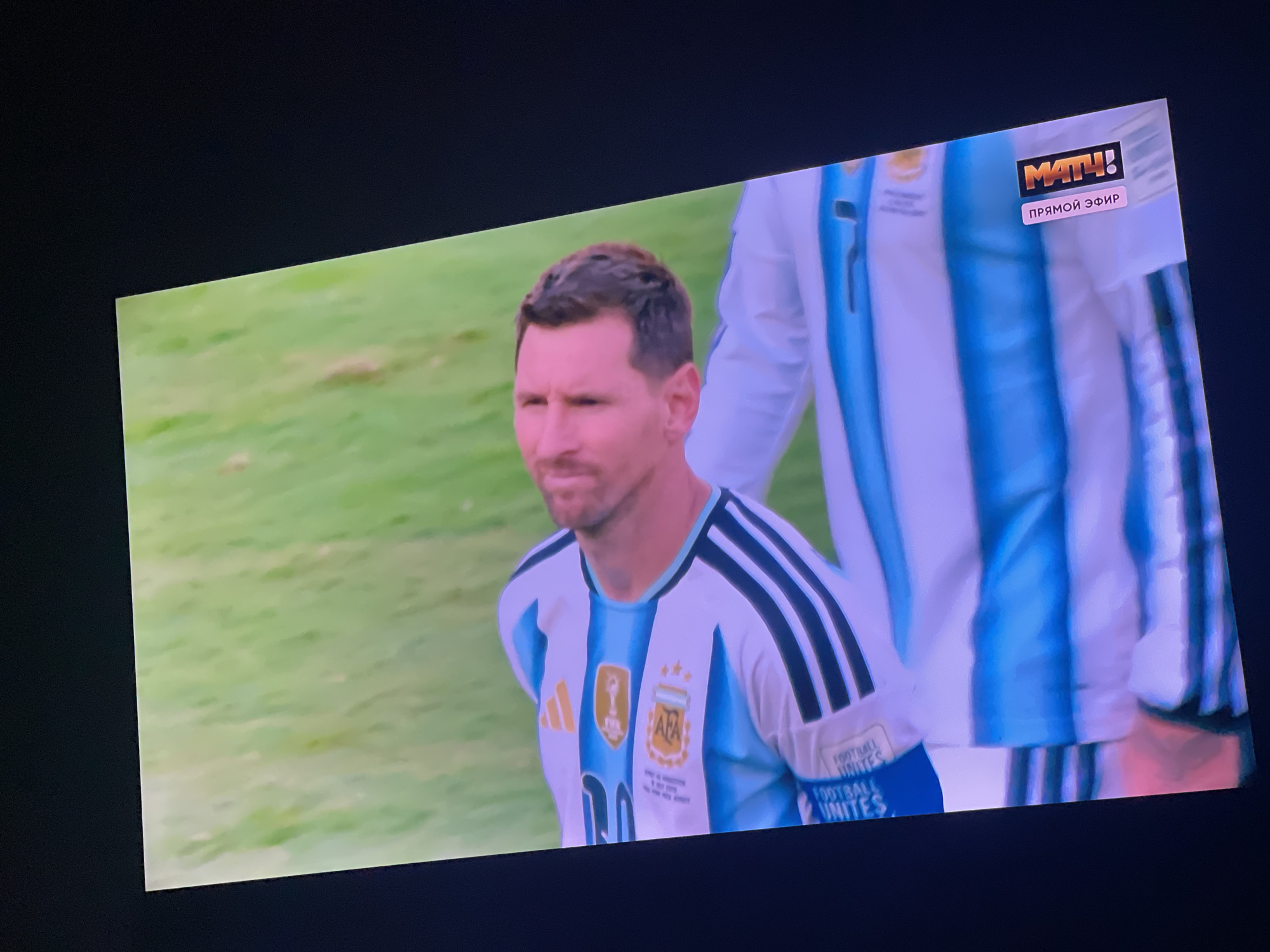 The image shows a close-up of Lionel Messi, a male person, wearing an Argentina national football team jersey. He is standing on a grassy field, likely a football pitch. Behind him, another person in a similar jersey is partially visible. In the upper right corner of the frame, a broadcast logo for "МАТЧ!" and the text "ПРЯМОЙ ЭФИР" (meaning "Live Broadcast" in Russian) are displayed. The scene appears to be a live broadcast of a football match. There is no visible indication of the city of Stary Oskol, Russia in the image.