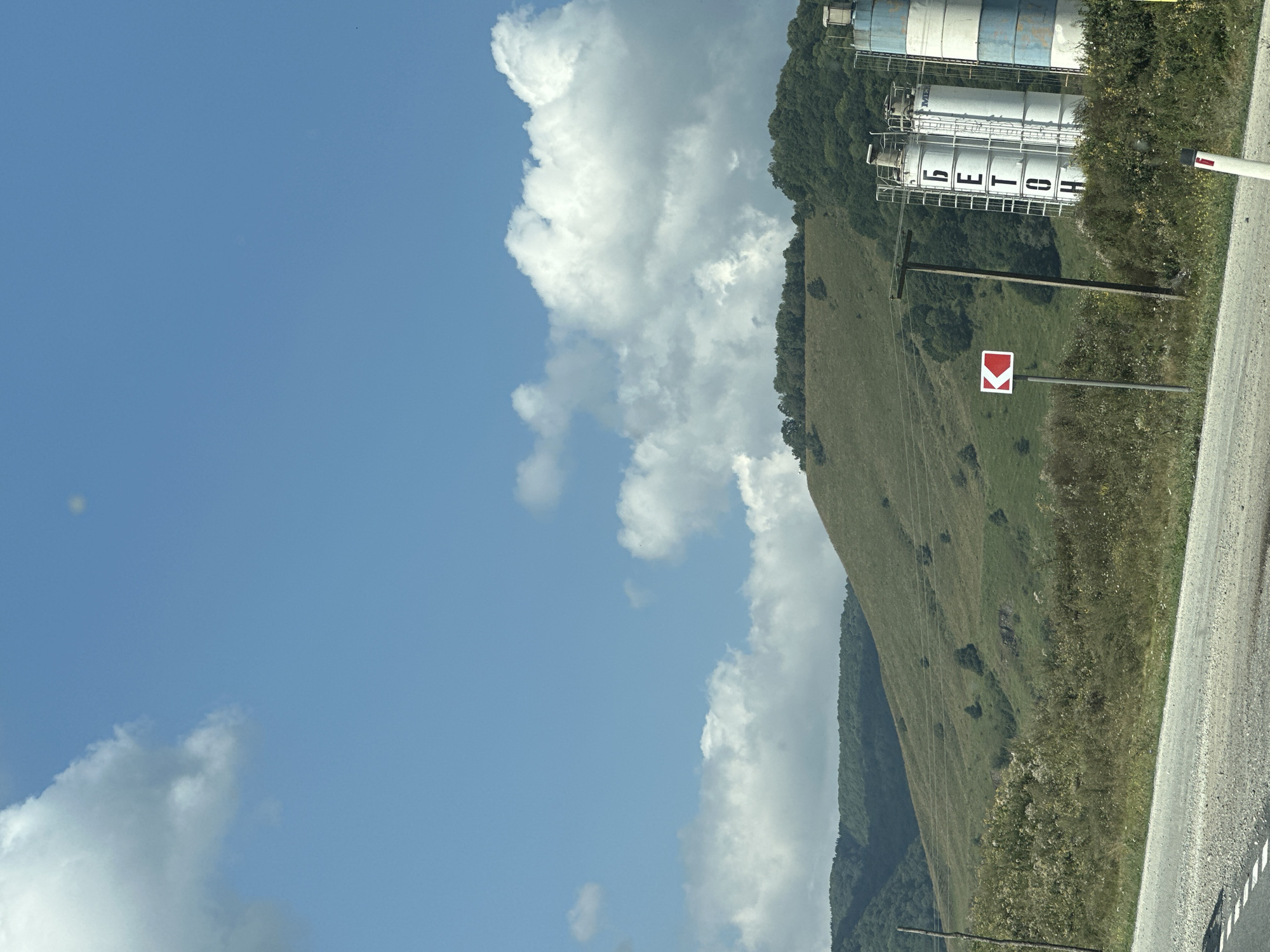 The image depicts a rural landscape with rolling green hills under a bright blue sky dotted with white clouds. In the foreground, a paved road with white markings is visible. To the right of the road, two industrial silos stand tall, one predominantly white with blue bands and Cyrillic lettering that reads "БЕТОН" (BETON), indicating concrete. A traffic sign with a red arrow pointing left is situated between the road and the silos. A wooden utility pole stands behind the silos. The background features lush green trees covering the hills. The scene appears to be located in Nizhnyaya Yermolovka, Russia, based on the provided location context. There are no people visible in the image.