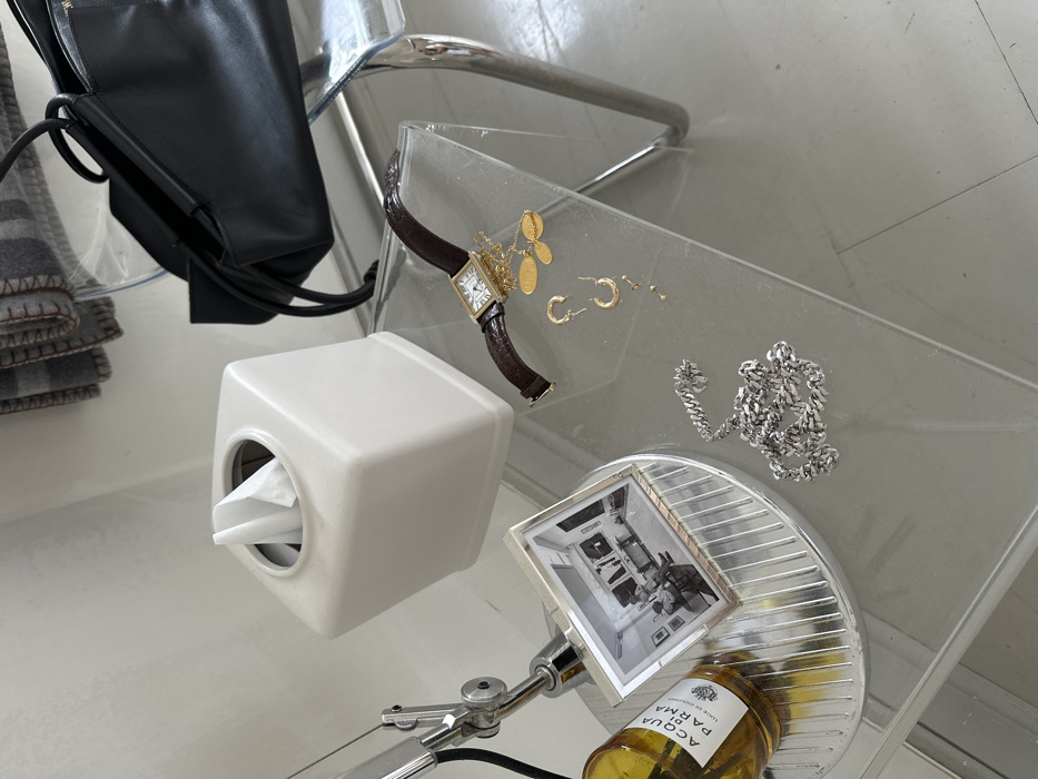 The image displays an indoor still life arrangement on a clear, rectangular acrylic or glass side table with curved chrome legs.
On the table, from left to right:
* An amber-colored glass bottle labeled "ACQUA DI PARMA" with a white label rests on a circular, fluted chrome coaster.
* Next to the bottle, a silver-framed black and white photograph depicts an interior room with artwork on the walls and furniture.
* A metallic, articulating lamp arm is partially visible on the far left.
* A white, square ceramic tissue box with white tissues protruding from its top opening.
* A rectangular-faced watch with Roman numerals and a dark brown, possibly crocodile-embossed, leather strap.
* A collection of gold-toned jewelry, including two hoop earrings, several small stud earrings, and a necklace featuring two flat, oval-shaped pendants.
* In the lower-left foreground, a silver-colored, curb-link chain is loosely coiled on the table surface.
In the background:
* A black leather handbag with twin top handles is positioned behind the watch.
* Behind the tissue box and handbag, a plain, light-colored wall is visible.
* Above the handbag and behind the tissue box, a section of grey patterned textile, possibly a blanket or throw, hangs against the wall.
The floor beneath the table is a light, uniform color, appearing off-white or very light grey. The image contains no identifiable landmarks, architectural features, or contextual elements to determine a specific location within the city of London, United Kingdom.