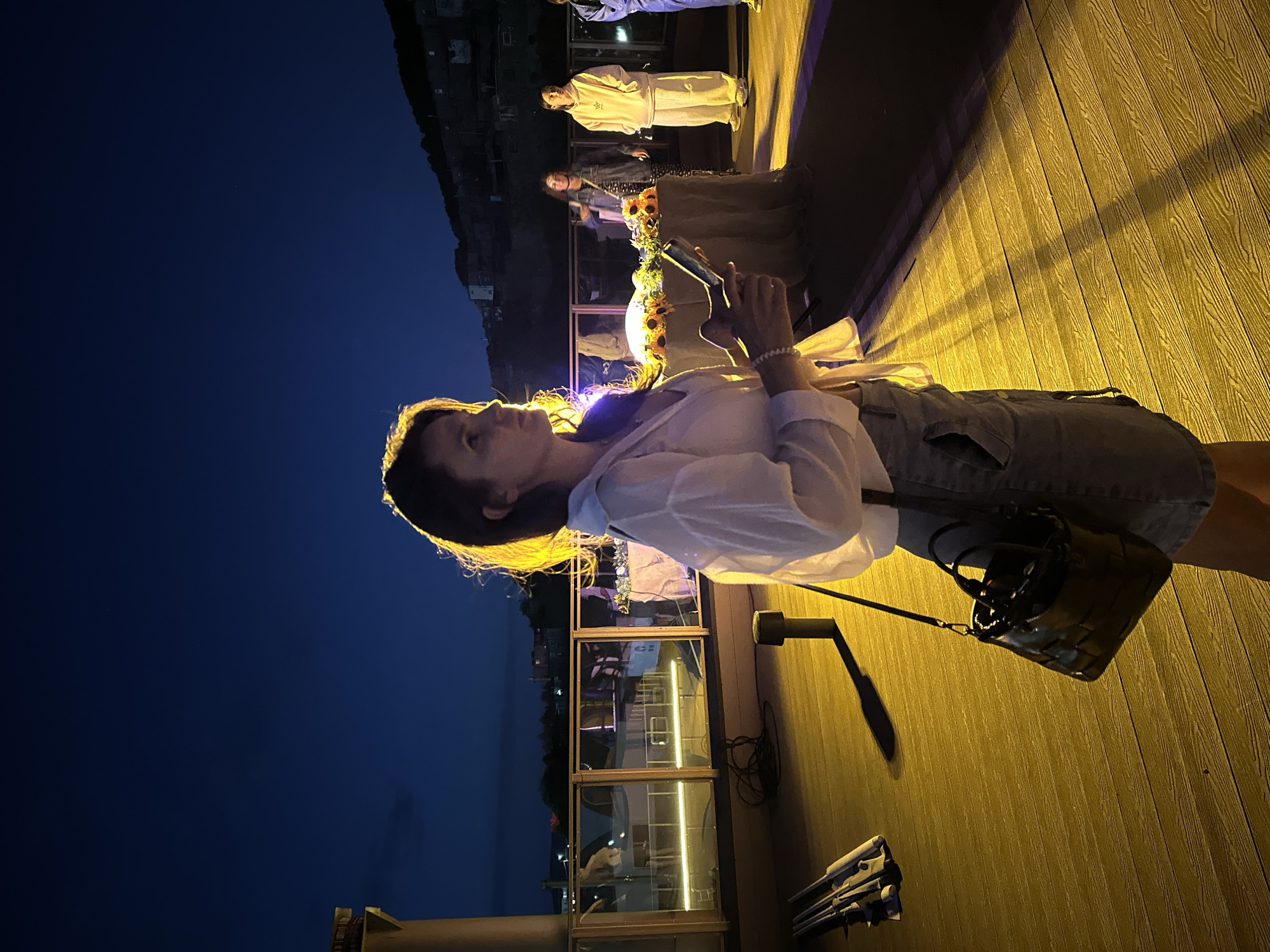 A woman stands on a wooden deck at dusk, illuminated by artificial light casting strong shadows. She is wearing a white open-collared shirt over a light-colored garment and a denim skirt. A dark handbag hangs from her shoulder, and she holds a smartphone in her right hand. In the background, a table is adorned with sunflowers. Several other people are visible in the distance, some standing and looking towards the table. Behind them, a body of water with boats and structures can be seen, with a hillside populated by buildings visible in the far distance under a dark blue sky. The scene appears to be an outdoor gathering or event. The architecture and hillside are consistent with a coastal city, potentially Petropavlovsk-Kamchatsky, Russia, known for its natural harbors and volcanic landscape.