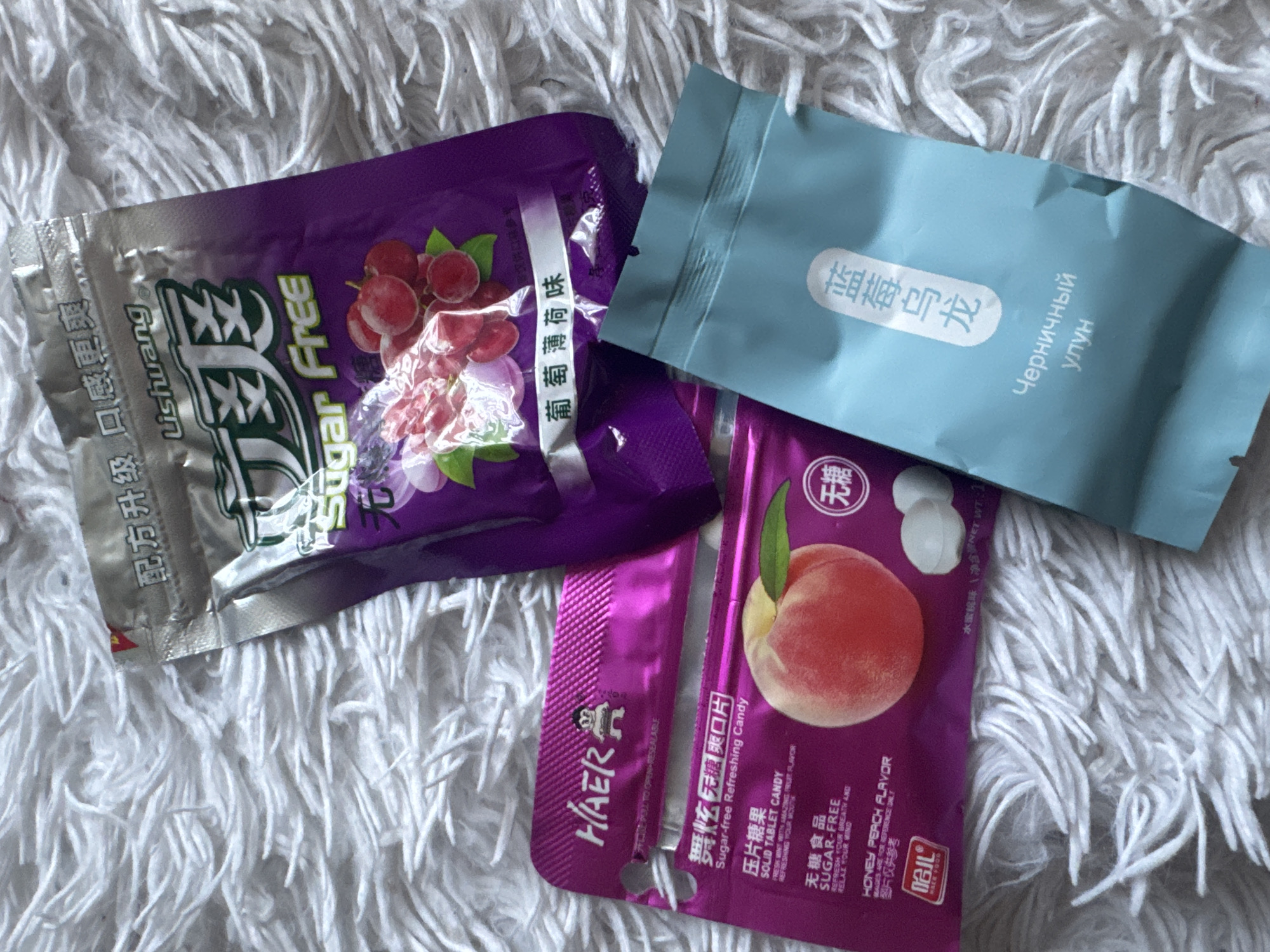 The image shows three unopened packages of sugar-free candy on a textured, off-white or light gray, fuzzy surface resembling a blanket or rug.
The first package is primarily purple with a silver section. It contains grape-flavored candy, as indicated by the grape illustrations and text "葡萄薄荷味". The brand name "Lishuang" is visible, and the package is labeled "Sugar Free".
The second package is light blue with text in Asian characters and Cyrillic, specifically "Черничный улун". Based on the Cyrillic text, this candy is likely blueberry oolong flavored.
The third package is magenta and features a peach illustration. Text on the package indicates it is "Sugar-free Refreshing Candy", "SOLID TABLET CANDY" and "HONEY PEACH FLAVOR." The brand name "HAER" is also visible.
There is no information in the image to suggest that the scene is located in Izhevsk, Russia.