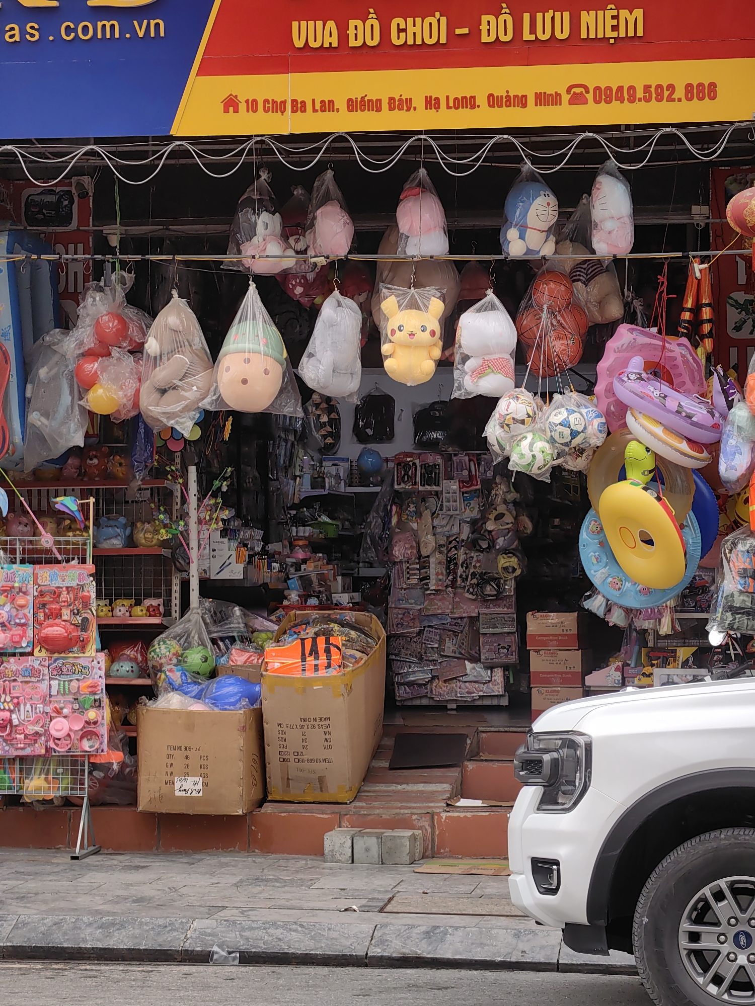 This eye-level, daylight shot captures the entrance of a vibrant toy and souvenir shop in Viet Hung Ward, Vietnam. The storefront is densely packed with merchandise, much of which is displayed hanging outside under an awning.
The subjects include a wide array of colorful toys and souvenirs. Prominently featured are plush toys like Pikachu, Doraemon, Hello Kitty, and various teddy bears, many individually wrapped in clear plastic bags and hanging from horizontal rods. Below these, several basketballs and soccer balls are also suspended. Inflatable swimming rings and pool toys, in various shapes and colors, hang towards the right side of the entrance.
Inside the shop, shelves are crammed with more toys, stationery, and other miscellaneous items. Two large cardboard boxes, partially obscuring the entrance, contain more merchandise. One box is labeled "ITEM NO.806-26", "QTY: 48 PCS", "G.W. 28 KGS", "N.W 25 KGS", and "MADE IN CHINA". Another box is labeled "ITEM NO.668-119" with similar shipping details and also "MADE IN CHINA". Boxes visible inside the shop are branded "KLONG" and "Composition Book".
The shop's red and yellow awning displays text: "VUA ĐỒ CHƠI – ĐỒ LƯU NIỆM" (King of Toys - Souvenirs). Below this, an address is printed: "10 Chợ Ba Lan, Giếng Đáy, Hạ Long. Quảng Ninh" and a phone number: "0949.592.886". Part of a blue sign above the awning reads "as.com.vn".
A white Ford vehicle, identifiable by its headlight, bumper, and tire with a Ford logo on the wheel hub, is parked partially in the foreground on the right, suggesting a street or parking area. The paved sidewalk and a curb with a small discarded plastic cup are visible in the immediate foreground. The scene depicts a lively commercial establishment, typical of a busy urban or town setting, under clear daylight conditions.
