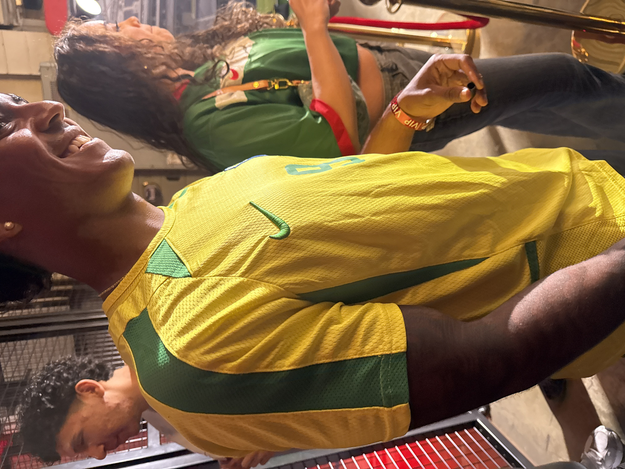 The image depicts two individuals. The person in the foreground is wearing a yellow and green athletic shirt with a Nike logo, smiling, and tilting their head back. The second person, in the background, has long curly hair and is wearing a green and red shirt. A "VIP" bracelet is visible on their wrist. They are standing near a gold-colored stanchion and red velvet rope. The location is suggested to be in Los Angeles, United States of America, but there are no specific indicators in the image confirming this.