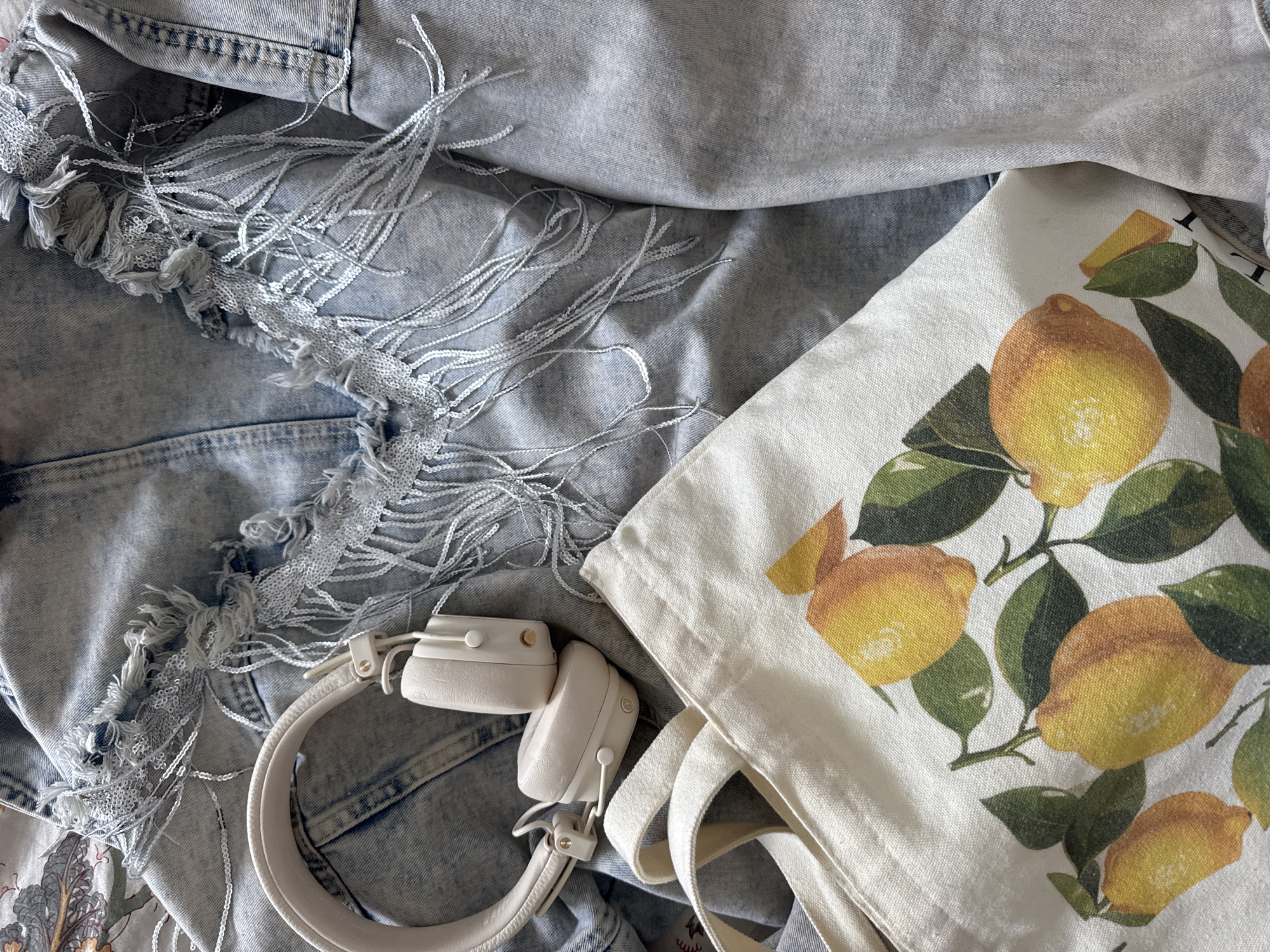 This is a close-up, overhead shot of a light blue denim jacket with distressed detailing and shimmering fringe, along with a light-colored canvas tote bag featuring a print of lemons and green leaves. A pair of light beige headphones rests on top of the jacket, partially obscuring the bag. The items are laid out on a surface that appears to be fabric, possibly a bedspread or blanket, with a subtle floral pattern visible in the background. The image was taken in Höhenkirchen-Siegertsbrunn, Germany. There are no people or actions depicted in the scene.