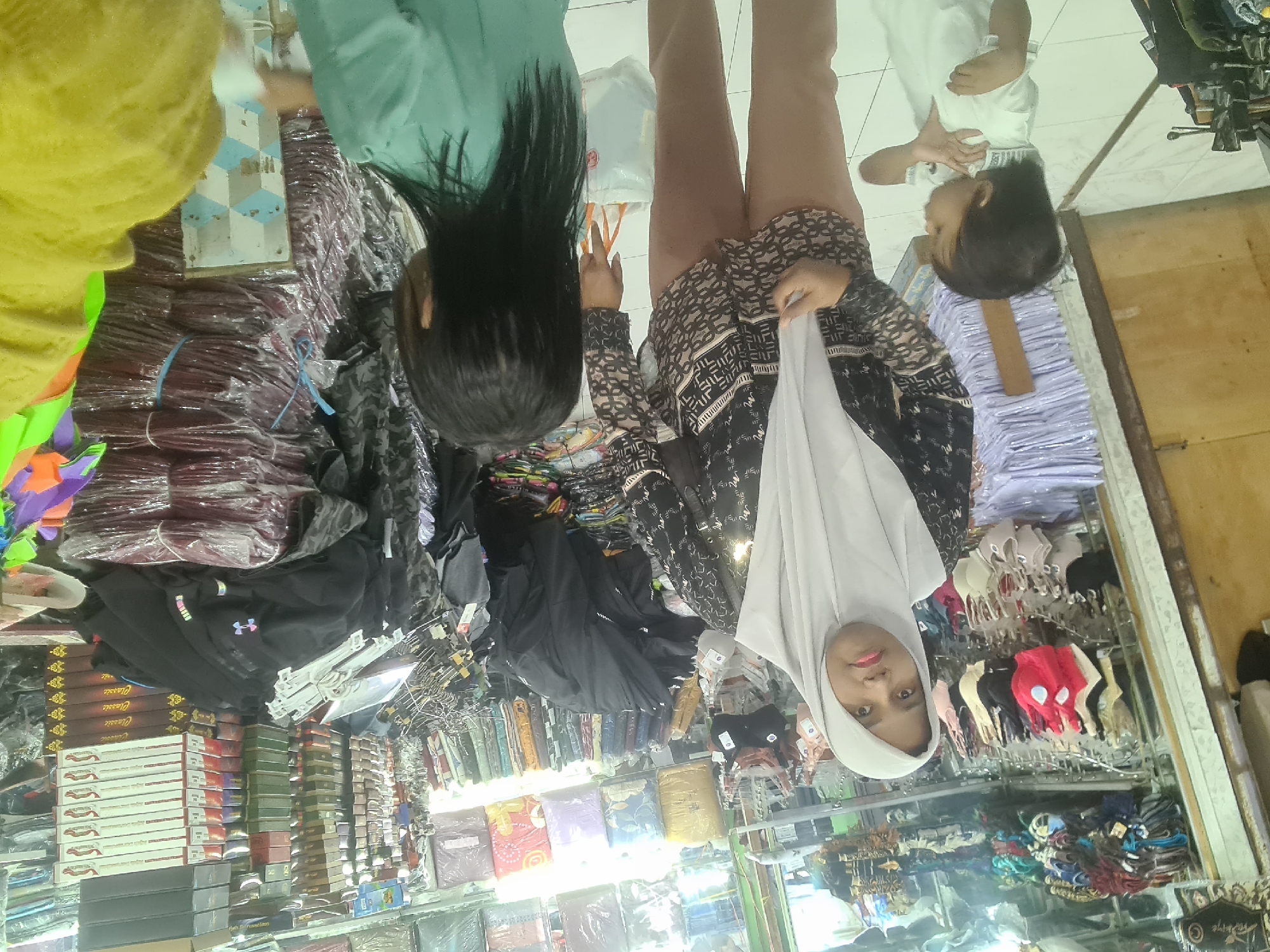 The image depicts the interior of a clothing store in Lhokseumawe, Indonesia. The store is filled with shelves and racks displaying various garments and folded merchandise. In the foreground, three individuals are visible. On the left, a young child in a white shirt is looking to their right. To the right of the child, a woman wearing a light grey hijab and a patterned black and white shirt stands facing the camera. Her lower body is clad in light brown trousers. She is holding a white plastic bag. To the right of the woman and facing away from the camera is another individual, likely a child, with dark, straight hair, wearing a light green top. The background is densely packed with goods, including stacks of fabric-wrapped items, boxes, and clothing racks filled with bras and other apparel. The store appears to be well-stocked, with a variety of colors and textures visible. The lighting suggests an indoor commercial environment.