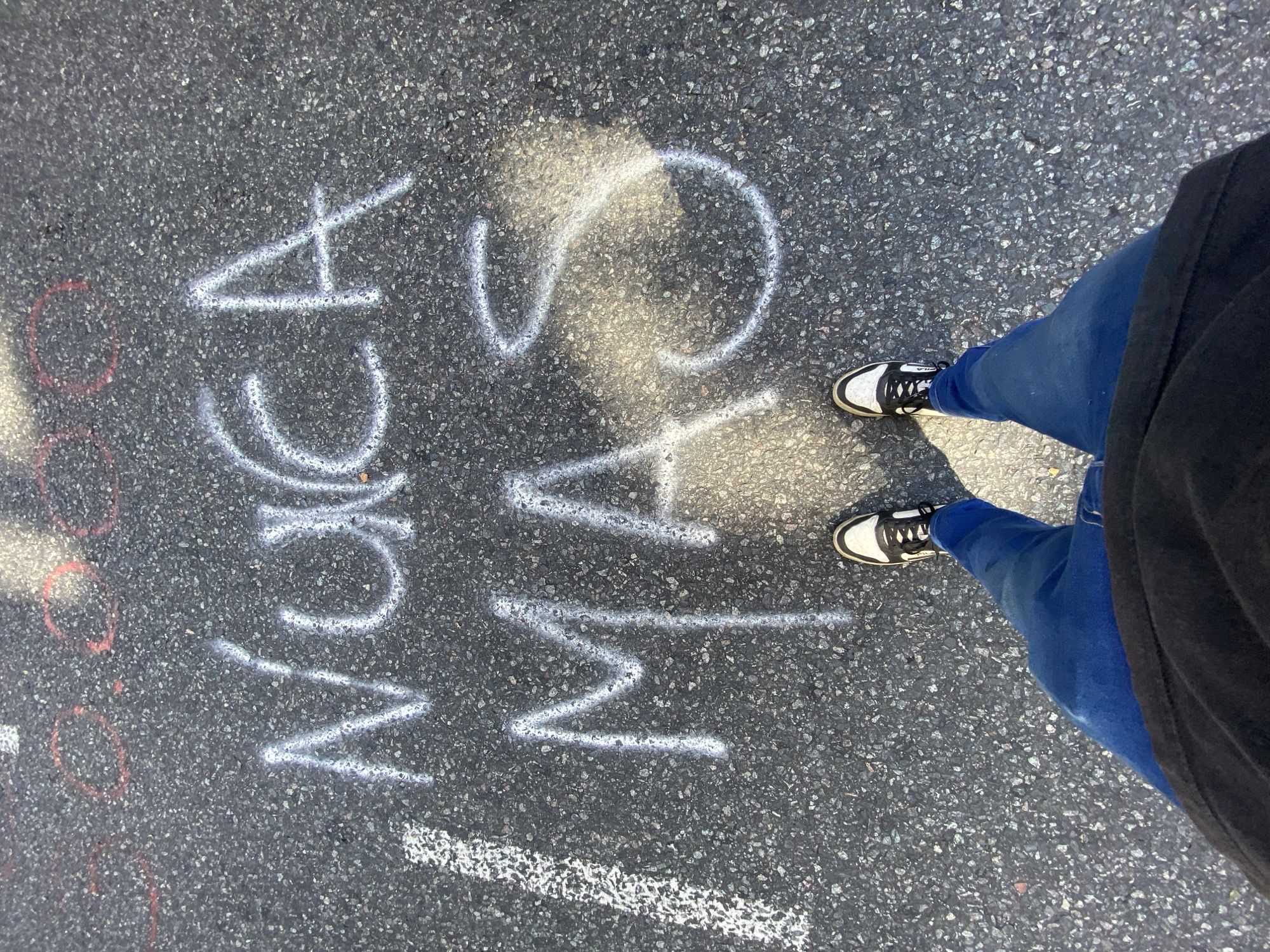 The image shows asphalt pavement with the word "MAS" written in large white letters, rotated 90 degrees clockwise. To the left of "MAS," the word "MCA" is written in similar style, rotated 90 degrees counter-clockwise. A person's legs and feet, wearing blue jeans and white/black sneakers, are positioned on the right side of the image, partially obscuring the word "MAS". The environment is outdoor, indicated by the pavement.
The exact location within Buenos Aires cannot be determined.