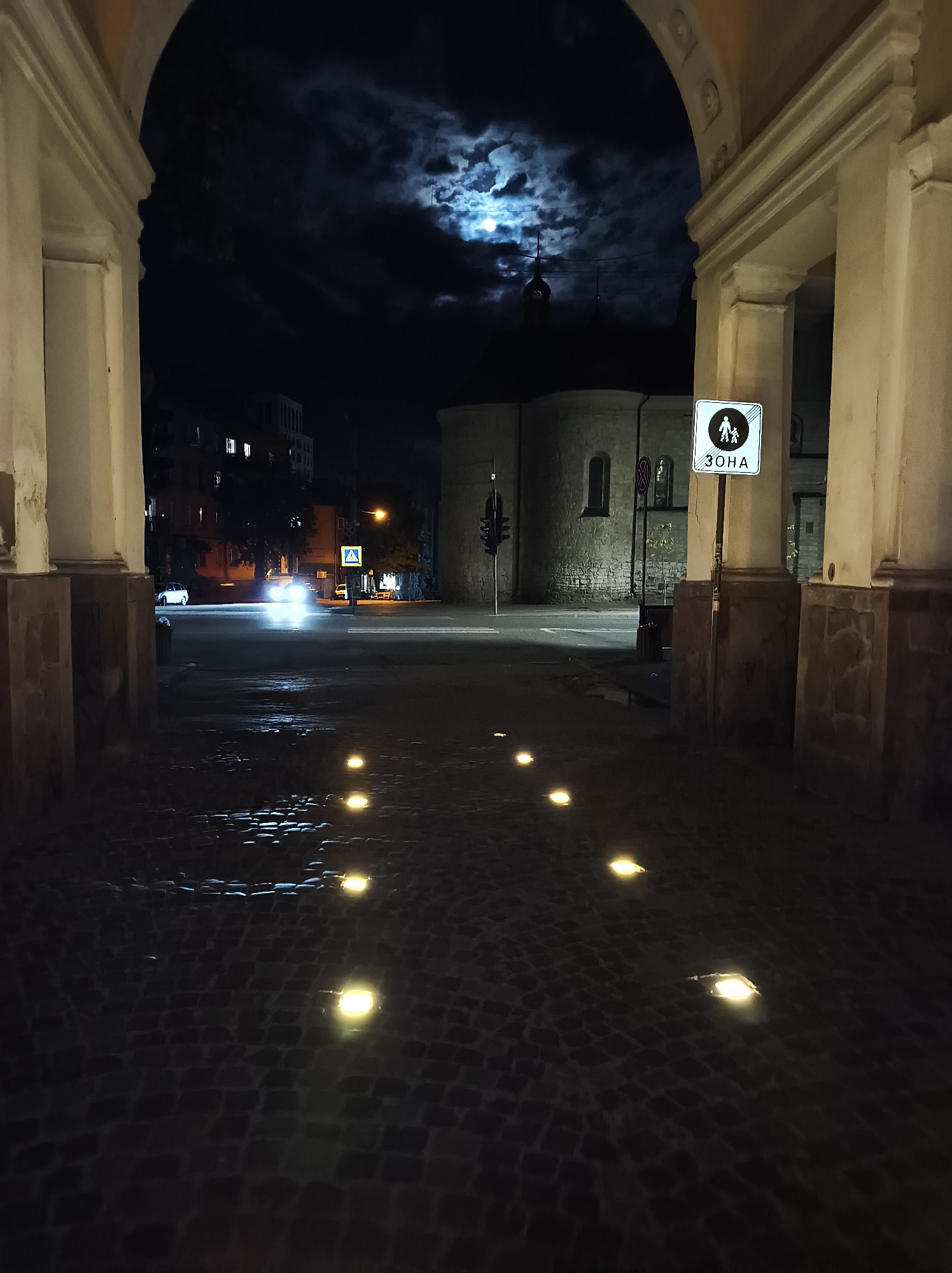 The image depicts a night scene in Ternopil, Ukraine, viewed through an archway. The archway is made of light-colored stone with architectural details. Beyond the archway, a street with a cobblestone surface is illuminated by a series of embedded lights. In the background, there's a building with a rounded roof, likely a church, visible under a cloudy night sky with a bright moon. A traffic light and a pedestrian zone sign in Cyrillic script (ЗОНА) are also visible on the right. The headlights of a car are seen approaching in the distance on the left.
