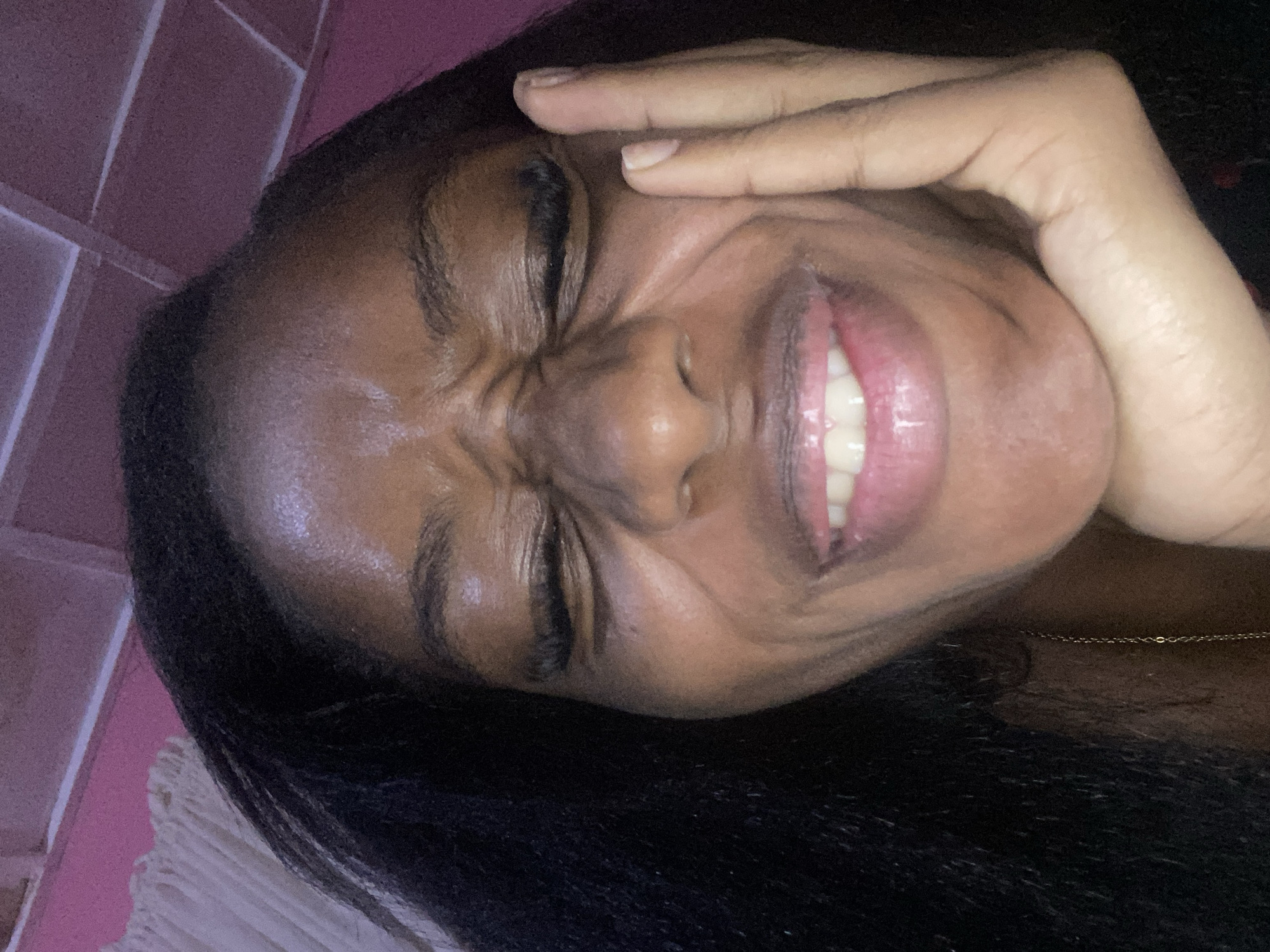 The image shows a close-up, overhead shot of a young Black woman's face, her eyes squeezed shut and her face contorted in what appears to be a laugh or grimace. She is resting her right hand on her cheek, and her dark hair frames her face. The background is out of focus, but appears to be an indoor setting with a pink-hued ceiling and a white curtain to the left. There are no discernible landmarks or visual cues to indicate the location is Jos, Nigeria.