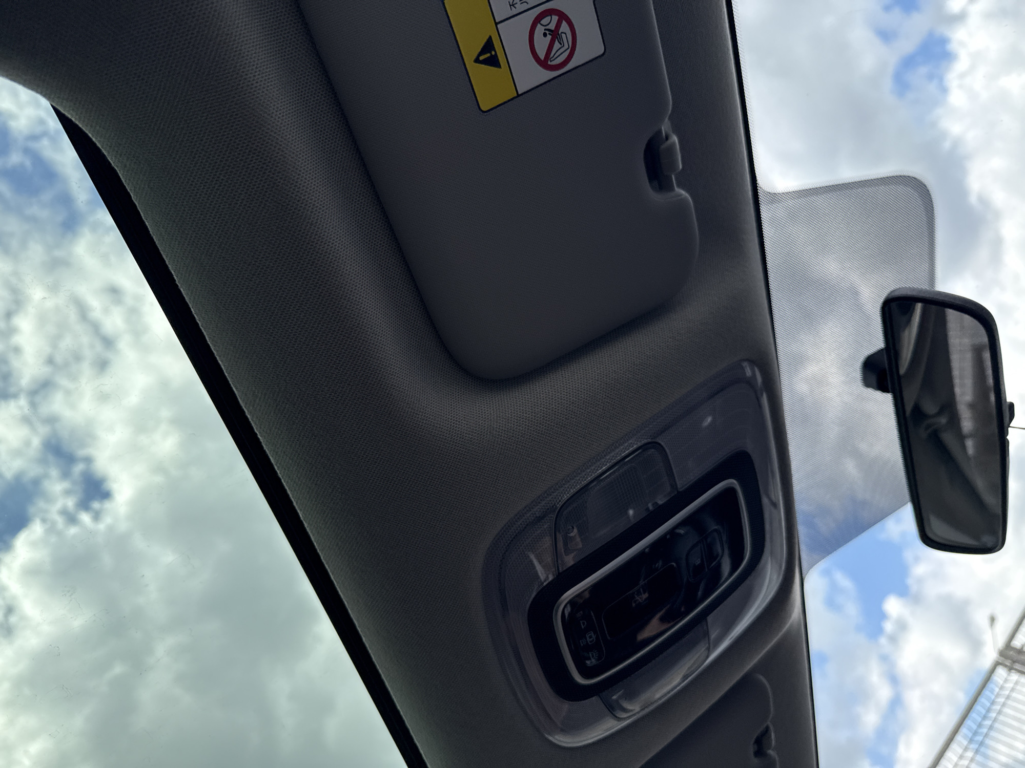 The image shows the interior of a vehicle, specifically the roof area. A gray sun visor is positioned in the upper left, with a yellow warning label attached. The car's interior lighting fixture is visible in the center, and a rearview mirror is mounted near the windshield on the right. The sky with clouds is visible through the windshield. No people are visible.