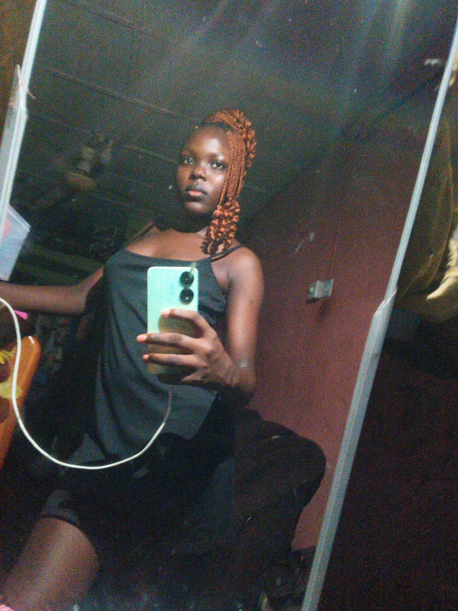 A young woman with braided hair takes a selfie in a dimly lit room, possibly a bedroom, in an apartment. She is holding a light green smartphone in her right hand, which is capturing her reflection in a large mirror. The woman is wearing a black camisole top. The background of the room is indistinct, but a wall with a reddish-brown hue and some objects on it are visible in the reflection. The lighting is poor, creating a grainy effect on the image. The overall impression is that of a casual, personal moment captured at home.