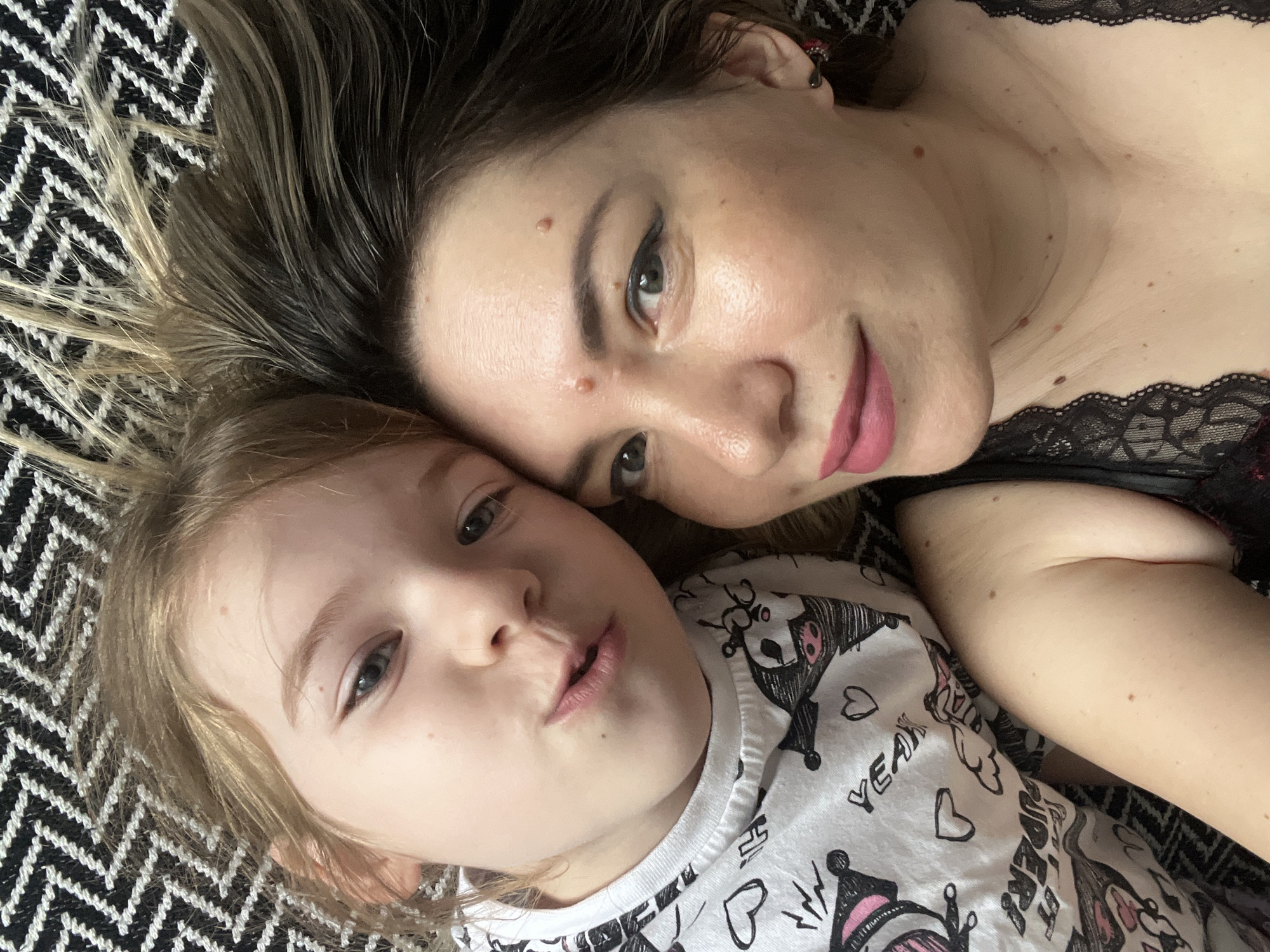 The media file shows a close-up selfie of a woman and a young child lying down on a black and white patterned surface, likely a blanket or rug. The woman is on the right, with her head tilted towards the child. She has dark hair with lighter streaks and is wearing black lace top. Her face shows a gentle smile and her eyes are looking towards the camera. The child is on the left, with blondish hair and is looking towards the camera with a playful expression, puckering their lips. The child is wearing a white t-shirt with a cartoon print featuring characters and text. The scene appears to be indoors, given the lighting and the close-up framing. The overall mood is intimate and affectionate, suggesting a moment of connection between the mother and child. The location context provided is Sochi, Russia. There is no discernible time of day or weather information present. The visible text on the child's shirt includes "YEAH" and some stylized lettering and drawings.
