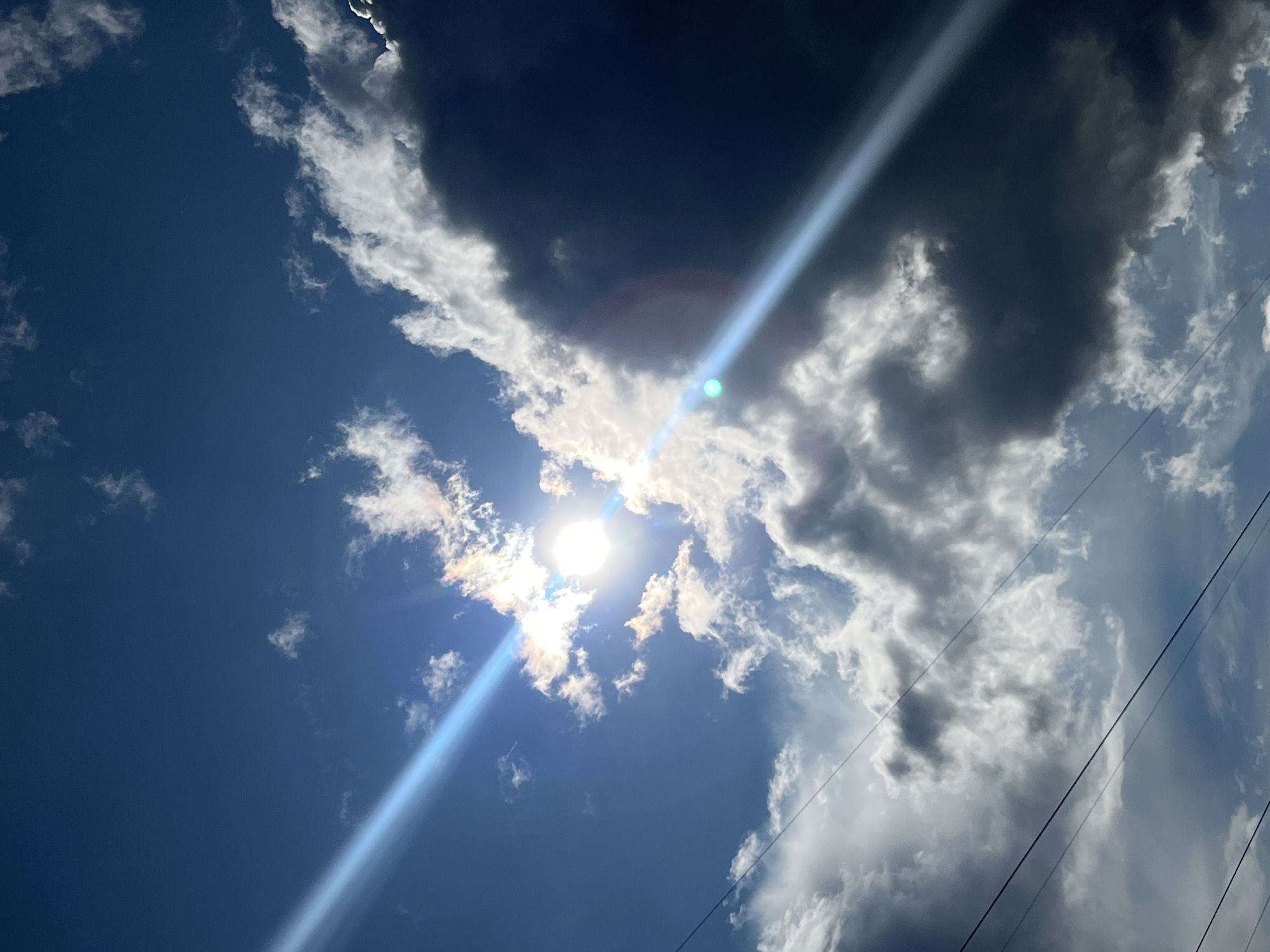 The image is a close-up, upward-facing view of a cloudy sky with the sun partially obscured by clouds. The sun appears bright and a sun flare is visible, creating a diagonal light beam across the frame. Some clouds are white and fluffy, while others are dark and ominous. There are also power lines visible in the lower portion of the image. The scene is characteristic of an outdoor environment, possibly in the city of Nizhny Tagil, Russia, given the context provided. No people or specific actions are depicted, only the natural environment and man-made infrastructure.