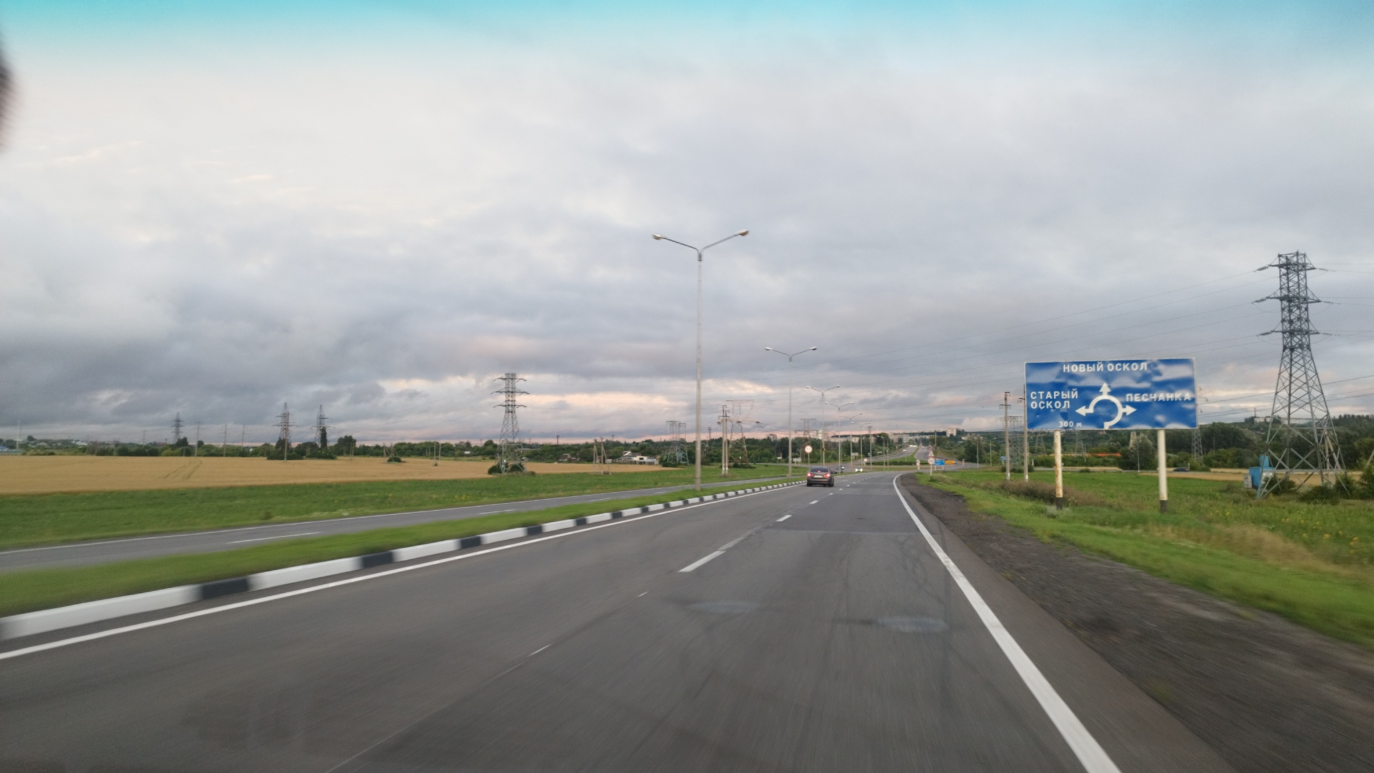 The image depicts a highway scene in Stary Oskol, Russia, likely captured during twilight due to the soft lighting and cloudy sky. In the foreground, a paved road with white lane markings stretches into the distance. The road is flanked by green grass and a white and black striped barrier. In the mid-ground, a single dark-colored car is visible on the road. On the right side of the road, a blue directional sign indicates "НОВЫЙ ОСКОЛ" (Novy Oskol) at the top, with "СТАРЫЙ ОСКОЛ" (Stary Oskol) and "ПЕСЧАНКА" (Peschanka) written below, along with a circular arrow symbol representing a roundabout and "300 м" (300 meters). Beyond the road and fields, several tall electrical towers and power lines are scattered across the landscape. The sky is filled with a mix of grey and white clouds, with a hint of light blue at the very top. The overall environment appears to be rural or semi-rural, with agricultural fields visible on the left.