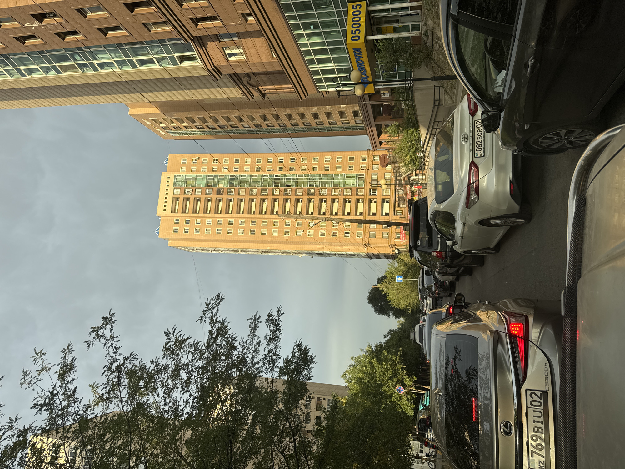 The image depicts a street scene in Almaty, Kazakhstan, likely during rush hour, with heavy traffic congestion. Several modern, multi-story buildings line the street, featuring beige and brown facades with numerous windows. In the foreground, a silver Lexus sedan with license plate "KZ 769BIU 02" is visible. Behind it, a white Toyota Camry with license plate "KZ 082BGR 02" is also in traffic. A dark-colored car occupies the right side of the frame. Trees with green foliage are present on the left side of the street, and traffic signs are visible, including one with a blue circle and a white pedestrian symbol. The sky is overcast with clouds. A yellow sign with blue lettering and the number "050005" is mounted on one of the buildings, indicating a post office or a similar service. The overall impression is one of urban development and the challenges of city transportation.