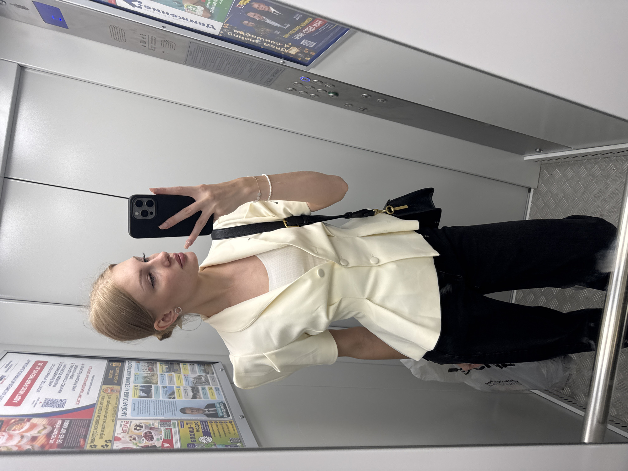A young woman with blonde hair tied back, wearing a cream-colored blazer over a white top and black pants, stands in an elevator and takes a selfie with her phone. She has a small black purse with a gold chain strap slung over her shoulder. The elevator is metallic and features an array of buttons and posters on the walls. The floor is made of a diamond-plate metal. The image appears to be taken in Perm, Russia, given the posters on the wall with Cyrillic text, which include advertisements and official notices.