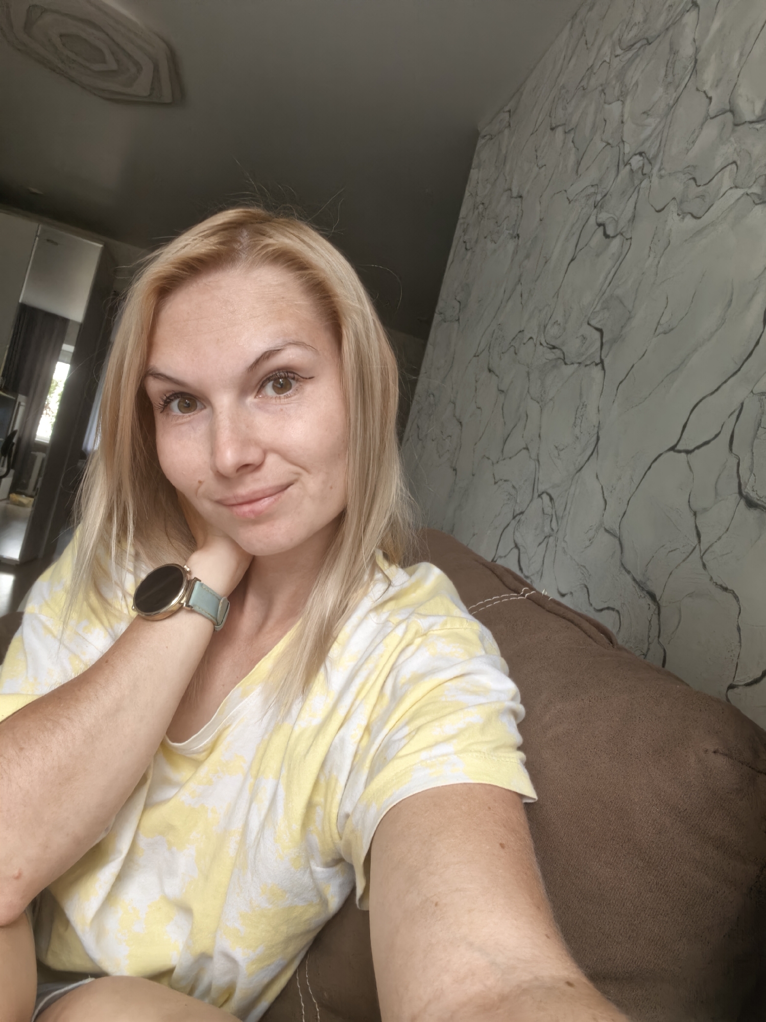 A woman with blonde hair and brown eyes is taking a selfie. She is wearing a yellow and white tie-dye t-shirt and a light blue smartwatch on her left wrist. She is sitting on a brown sofa, and behind her is a wall with a gray marble-like pattern. The image appears to have been taken indoors. There is no indication of the city of Sterlitamak, Russia in the image.