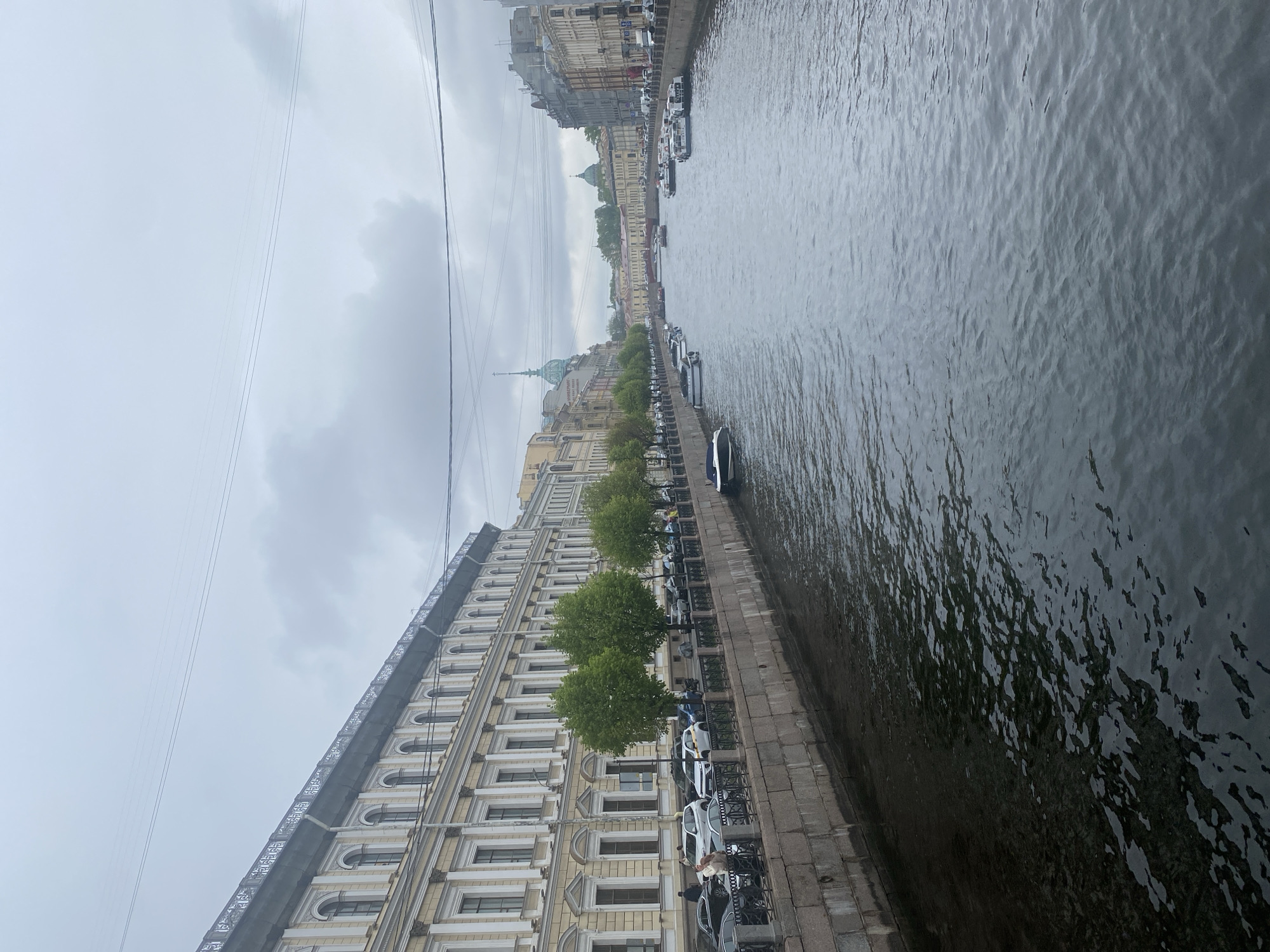 The image depicts a cityscape alongside a body of water.
Buildings with architectural details are visible, lining the embankment. Cars and trees are present along the embankment. Several boats are moored along the embankment. The water has a dark, reflective surface. The sky is overcast.
The scene is located in Saint Petersburg, Russia.