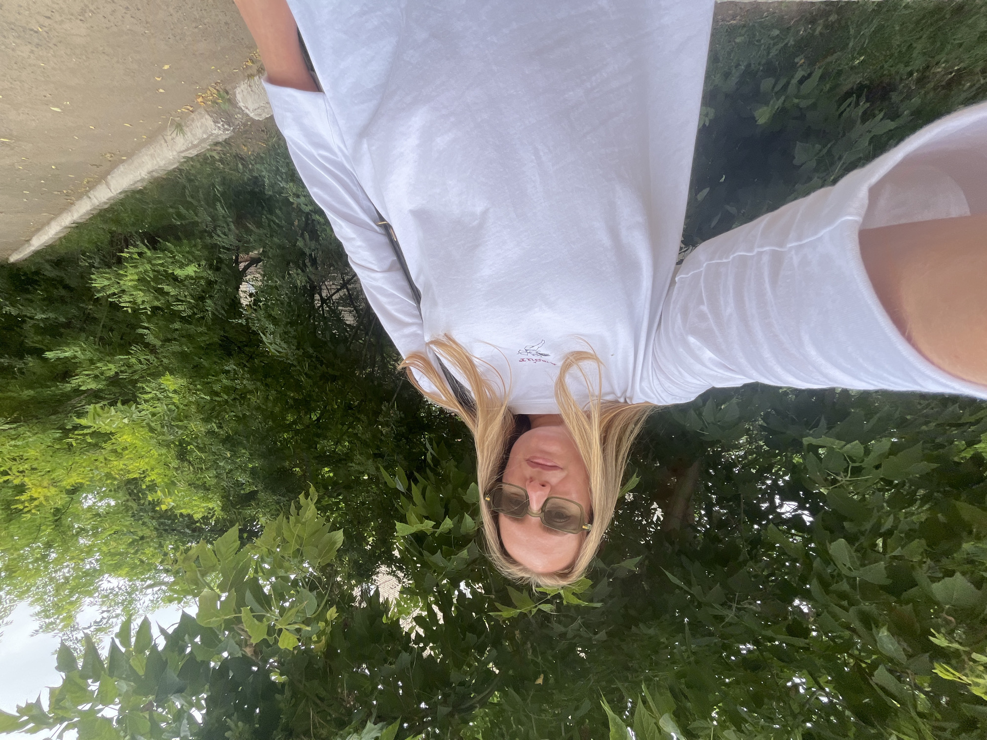 The image shows a blonde woman with sunglasses, wearing a white long-sleeved shirt. She is outdoors, with greenery surrounding her, suggesting a park or garden setting. The image is a selfie, taken from a low angle. The woman appears to be posing for the camera, with a neutral expression. The time of day is likely daytime, with bright lighting suggesting sunshine. Some text is visible on the woman's shirt, but it is too small and blurry to read. The overall impression is a casual, outdoor portrait. The context provided indicates the location is Chaikovsky, Russia.