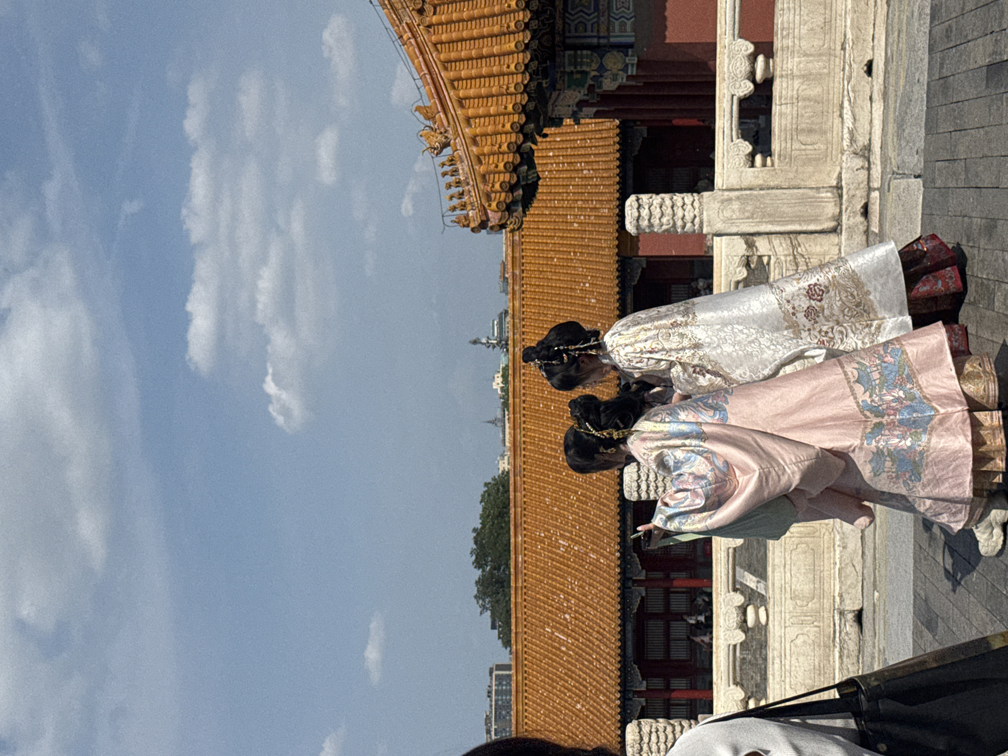 Three individuals, viewed from behind, are walking on a paved pathway in Dongcheng District, China. They are dressed in traditional Chinese attire with elaborate patterns. The person in the center wears a pink robe adorned with blue and green floral motifs, while the person to their right is in a white robe with intricate gold embroidery. A third person, partially visible on the left side of the frame, is wearing a light-colored top and carrying a dark tote bag.
Behind the individuals is a stone railing and a building with a distinctive orange tiled roof and red walls, characteristic of traditional Chinese architecture. In the background, there are more buildings, trees, and a blue sky with scattered white clouds. The overall scene suggests a visit to a historical site or cultural attraction.