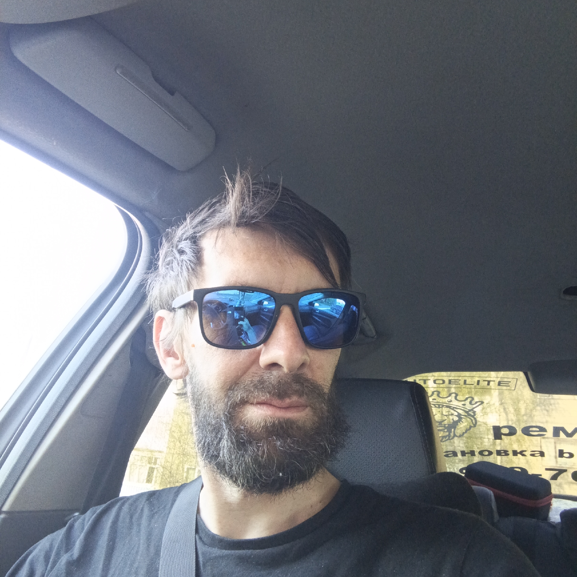 A man with a beard and sunglasses sits in the driver's seat of a car. He is wearing a black t-shirt and a seatbelt. The car is in Марковский, Russia, as indicated by a sticker on the back window with Russian text, including the words "Автоэлита Ремонтовка". The sunglasses reflect the car's interior and the person holding the camera. The hair is messy.