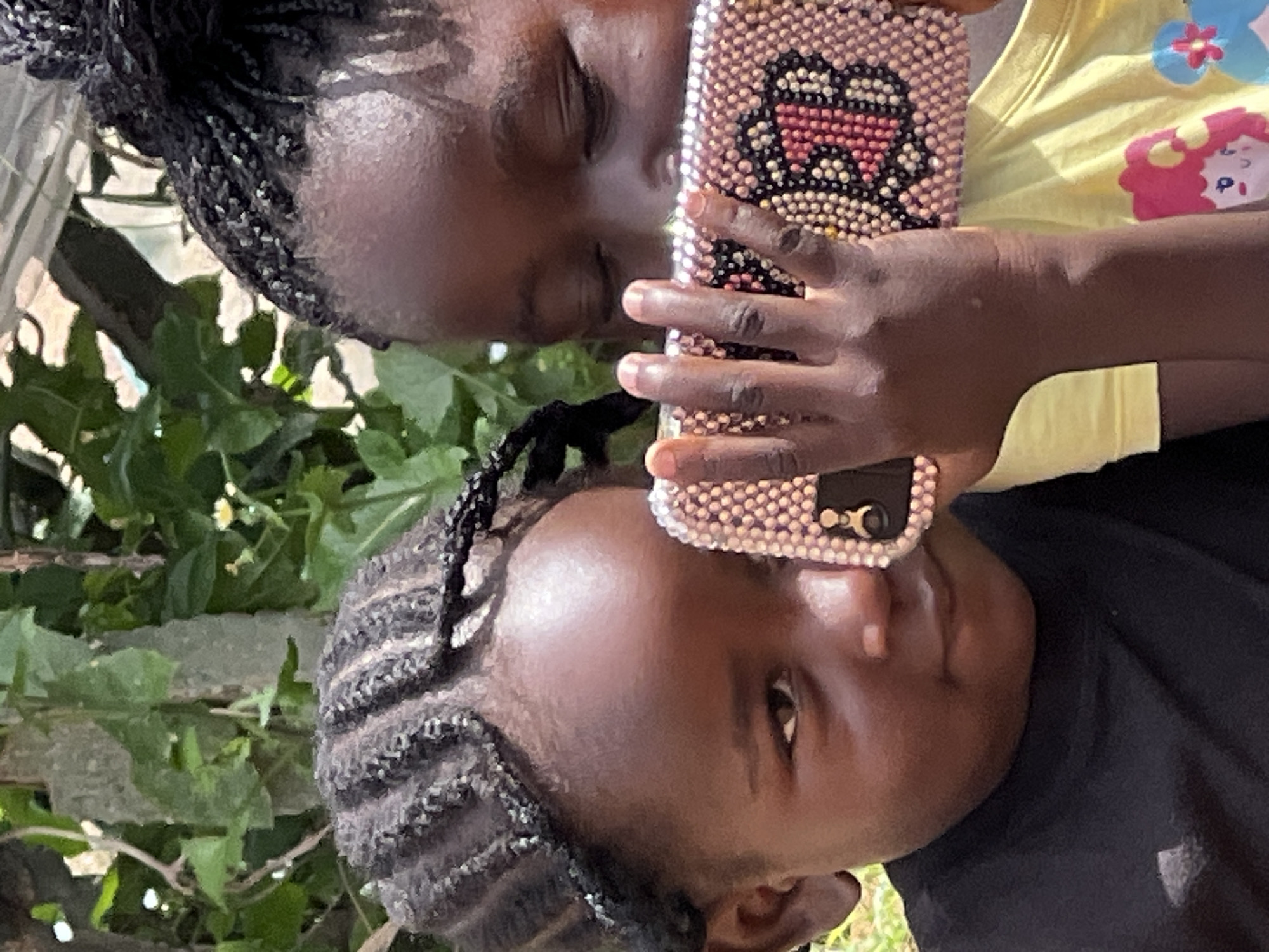 This is a close-up, medium shot of two young Black girls in an outdoor, natural setting. The girl on the left is facing forward, her face partially obscured by a mobile phone held by the girl on the right. Her dark, braided hair is visible on her forehead, and she is wearing a black t-shirt. Her gaze is directed slightly downward, towards the phone.
The girl on the right is mostly obscured by the phone, which she is holding horizontally with both hands. The phone case is decorated with numerous small, sparkling rhinestones in a pattern that includes what appears to be a large letter "B" in red and yellow. Her dark, braided hair is pulled back. She is wearing a bright yellow t-shirt with colorful cartoon characters on it.
The background consists of lush green foliage and branches, suggesting a garden or a wooded area. The lighting indicates it is daytime, with sunlight filtering through the leaves. There are no specific landmarks visible that would pinpoint the exact location within Jos, Nigeria, but the environment suggests a warm, possibly tropical or subtropical climate. The overall impression is one of childhood engagement with technology amidst nature.