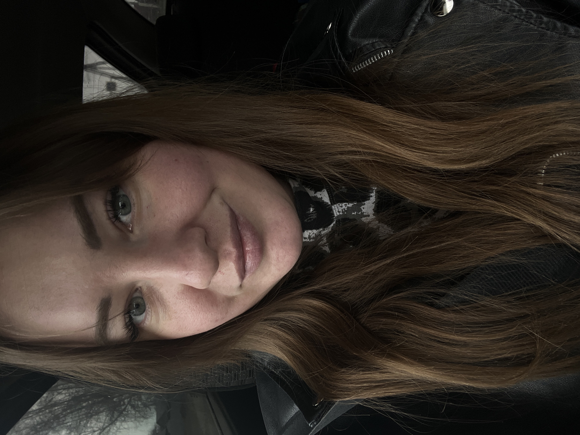 A close-up portrait of a light-skinned woman with long brown hair, wearing a black leather jacket and a black and white patterned turtleneck. The photo appears to have been taken inside a car, judging by the window frame and blurred background visible in the upper left. It is not possible to confirm the location as Chaikovsky, Russia from the image alone.