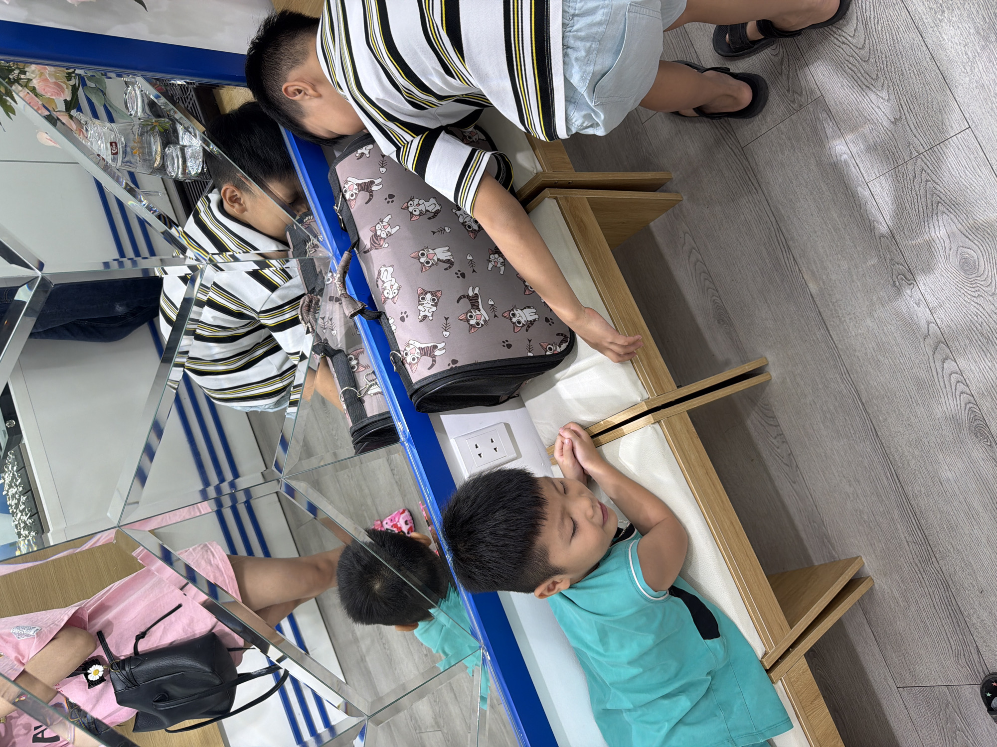 A child lies asleep on a bench, next to a backpack. A second person is leaning over the child and backpack. The image is primarily a reflection in a wall of mirrors. The setting appears to be indoors, with light-colored wooden flooring. No information indicates the image was taken in Đà Nẵng, Vietnam.