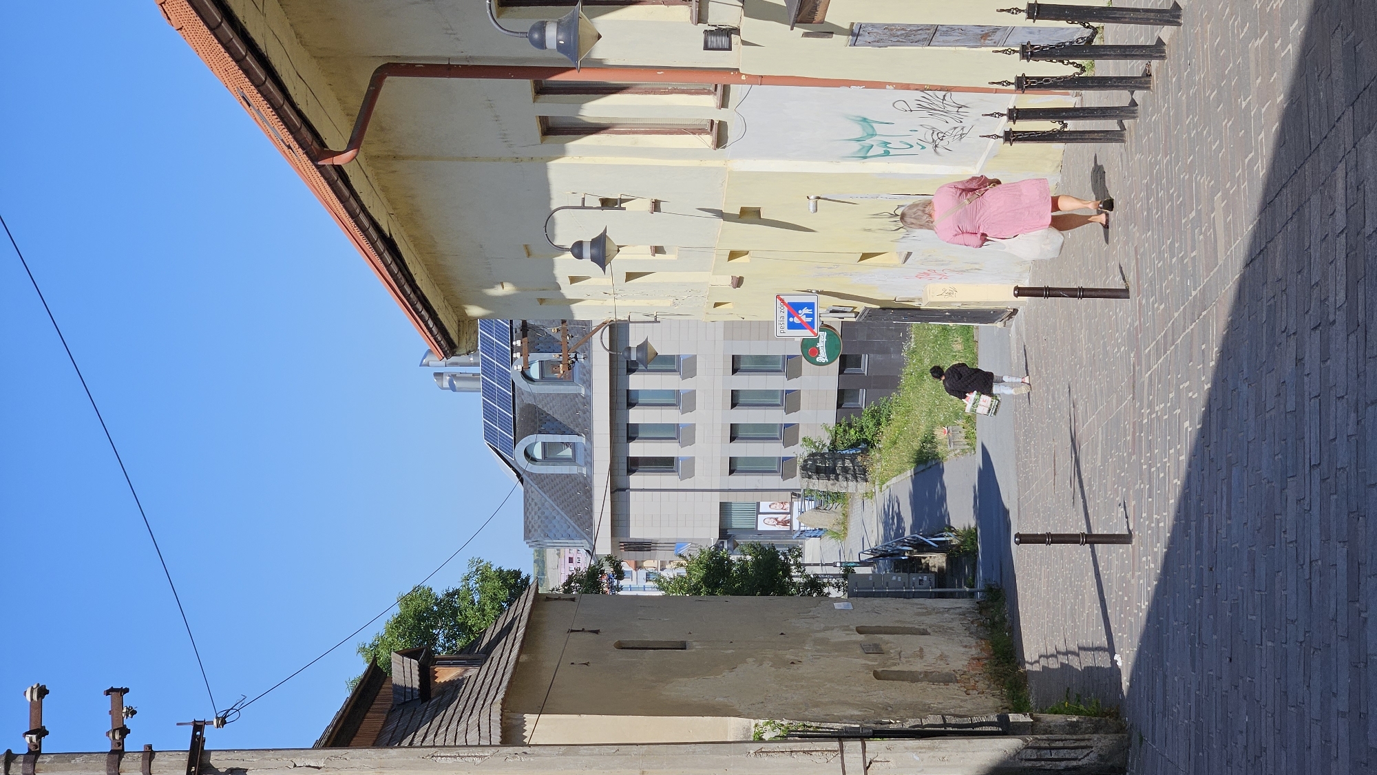 The image depicts a steep, cobbled alleyway in Prešov, Slovakia, with buildings on either side. In the foreground, two women are walking away from the camera up the alley. The woman on the right wears a pink dress and carries a white bag. The woman on the left wears a dark jacket and light pants, also carrying a white bag. The alleyway is lined with dark bollards and a metal chain fence on the right.
Behind the women, the alley continues uphill, with a paved walkway branching off to the left. Various buildings are visible in the background, including a modern building with solar panels on its roof and an older building with a wooden roof and dormer window. Signs are visible on the right side of the alley, including a pedestrian zone sign. The sky is clear and blue. The sunlight casts long shadows across the cobbled path. The overall scene suggests a sunny day in a historic urban environment.