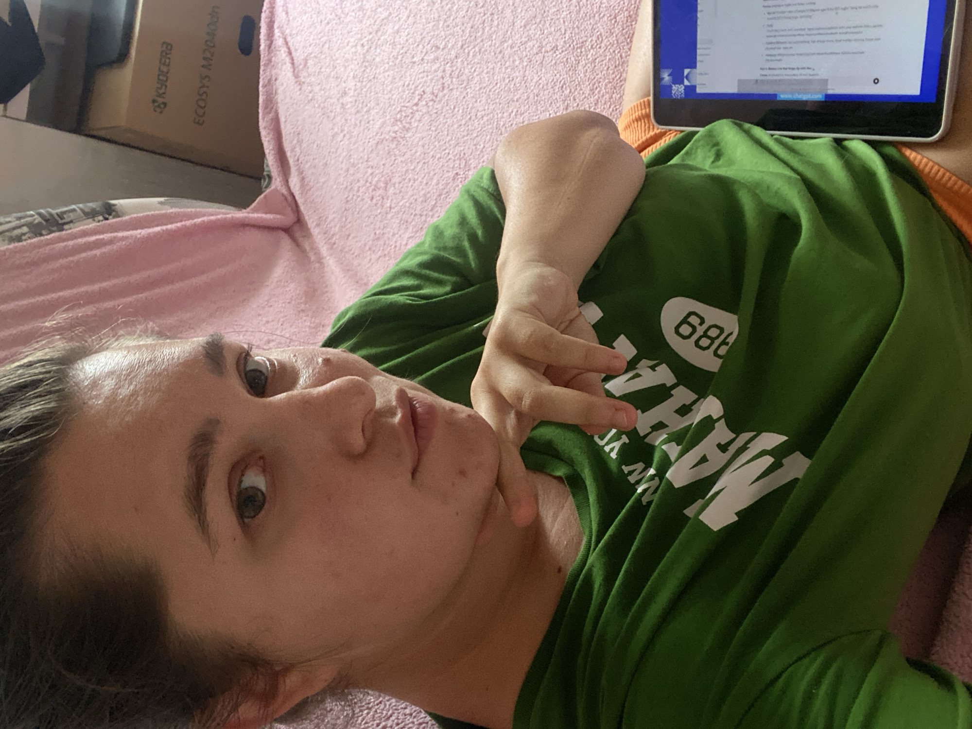 A woman is lying on a pink bed, holding a tablet. She is wearing a green t-shirt with the words "NEW YORK MATH" and "1989" printed on it in white. Her right hand is resting on her chin, and her left arm is bent at the elbow, resting on her chest. The tablet, positioned on her right side, displays text on its screen. In the background, a cardboard box with "KYOCERA" and "ECOSYS M2040dn" printed on it is visible. The scene appears to be indoors, likely in a bedroom. The provided metadata does not indicate the location of the image in the city of Perm, Russia.