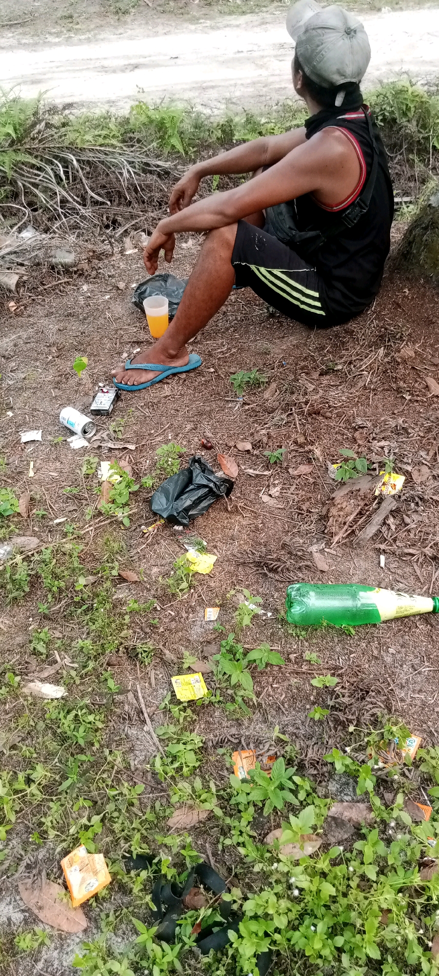 In the city of Unknown, Indonesia, a person sits on the ground under the shade of a tree. The individual is wearing a black tank top, black shorts with green stripes, and a light-colored cap. They are seated with their legs crossed, facing away from the camera, and appear to be resting. The surrounding environment is littered with various items, including plastic bottles, cans, cigarette packs, and plastic bags. The ground is covered in dry leaves, dirt, and patches of green vegetation. The overall scene suggests an outdoor, possibly undeveloped area, with evidence of human activity and discarded waste.