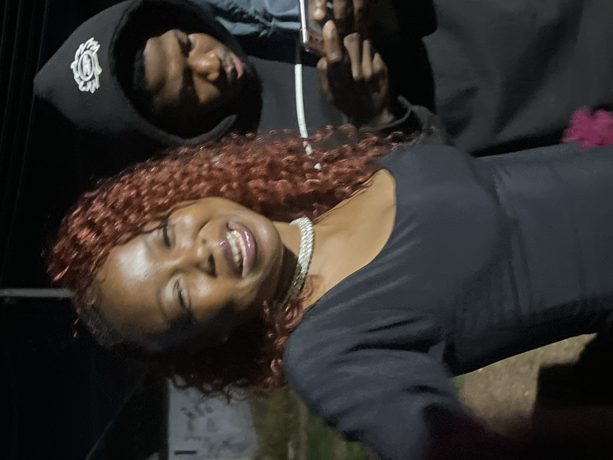 An outdoor, low-light, waist-up shot features a Black woman with reddish, curly hair smiling at the camera, her eyes closed. She is wearing a black top and a silver necklace. Behind her to the right, a Black man wearing a black hoodie with a white emblem is looking down at his phone, his face partially obscured by the hood. The background is dark, with some indistinct structures and foliage visible. The image appears to have been taken at night. The location is specified as Durimi, Nigeria, though this cannot be independently verified from the image itself.