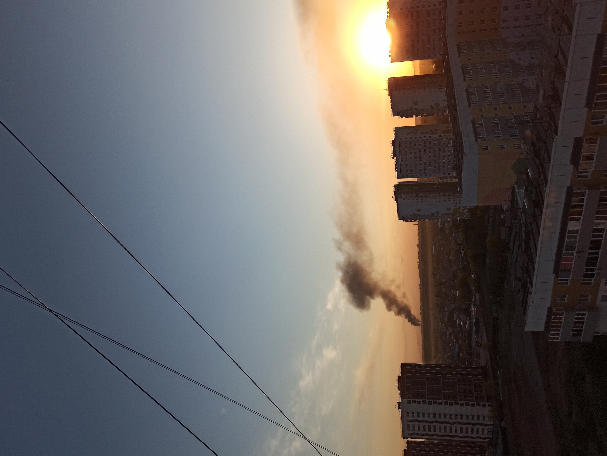 Here is a description of the image:
The image depicts a cityscape at sunset, likely in Кондратово, Russia, based on the presence of typical urban buildings in the area. Multiple high-rise buildings dominate the skyline, illuminated by the setting sun, which casts a warm orange glow across the scene. The sky is a gradient, transitioning from a dark blue in the upper left to a lighter orange near the horizon. A plume of dark smoke rises prominently from an area beyond the buildings, suggesting a fire or industrial activity. Power lines are visible in the lower left of the frame. The image appears to have been taken from an elevated perspective, offering a panoramic view of the city and the surrounding environment.