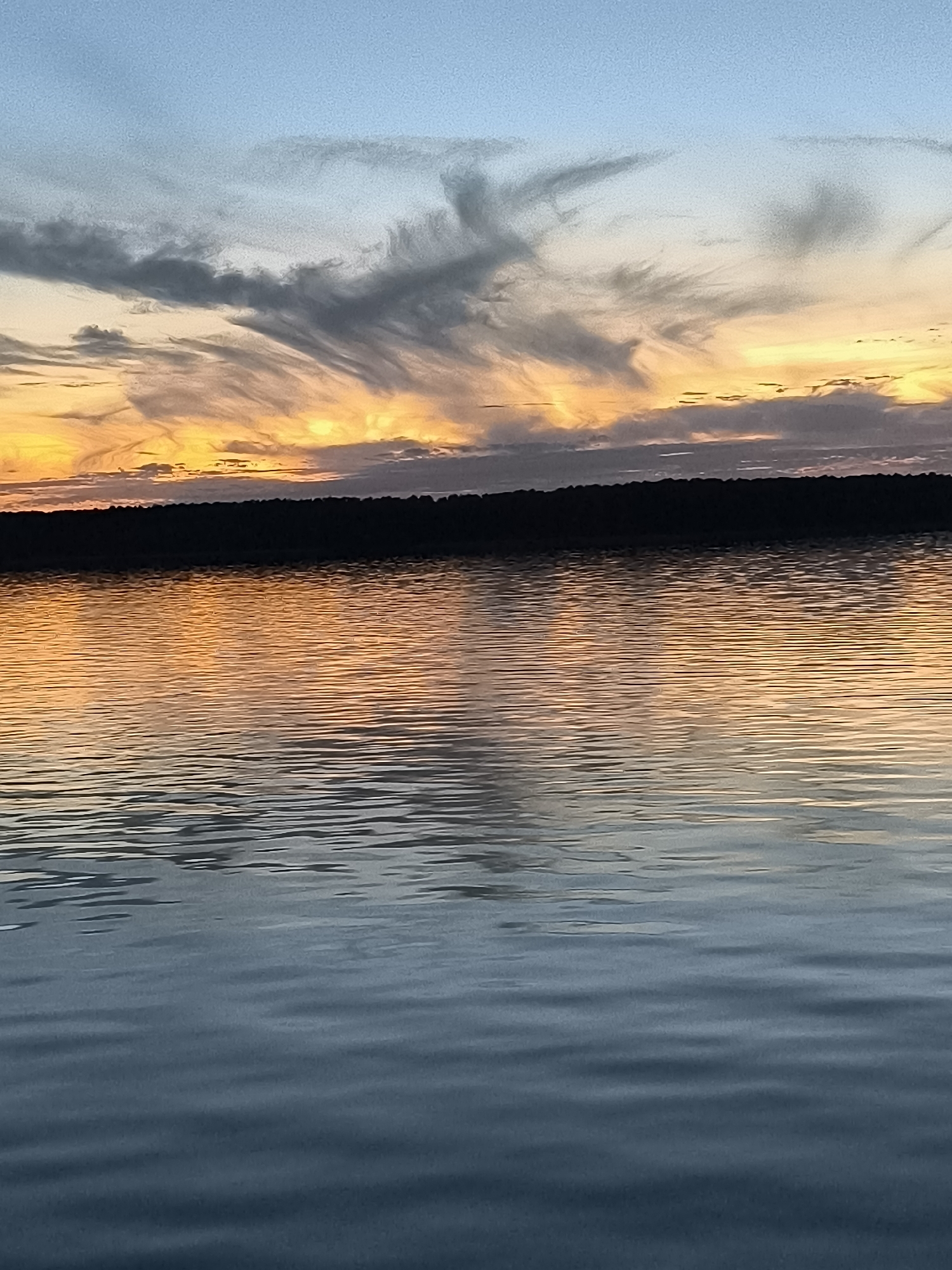 A serene sunset over the Curonian Lagoon in Neringa, Lithuania. The sky is a gradient of pale blue at the top, transitioning to fiery orange and yellow hues near the horizon, painted with wispy, streaky clouds. A dark, silhouetted treeline forms the distant shore. The water of the lagoon is calm, with gentle ripples reflecting the vibrant colors of the sunset, creating a mirrored effect of the sky. The foreground of the water is a deeper blue-grey, indicating the transition from dusk. There are no people or objects visible in the scene, emphasizing the natural beauty of the landscape at twilight.
