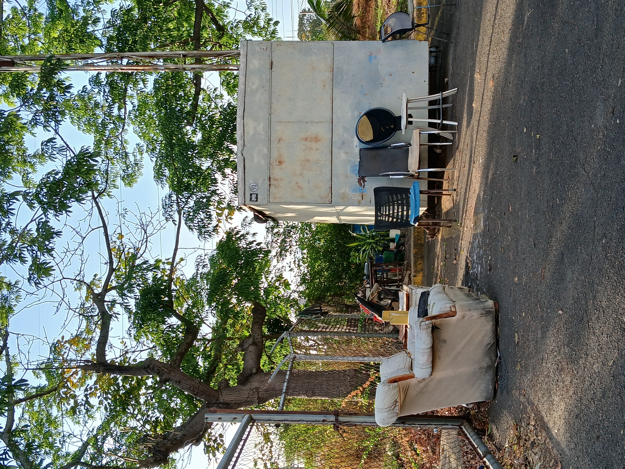 The image depicts discarded furniture, including a sofa and multiple chairs, placed beside a large metal container or structure on a paved street. A chain-link fence runs parallel to the street. Trees are present on both sides, partially obscuring the sky. The location is in an unidentified part of Ciudad Guayana, Venezuela. There are no people visible in the image. The scene presents an impression of abandoned or disposed-of items.