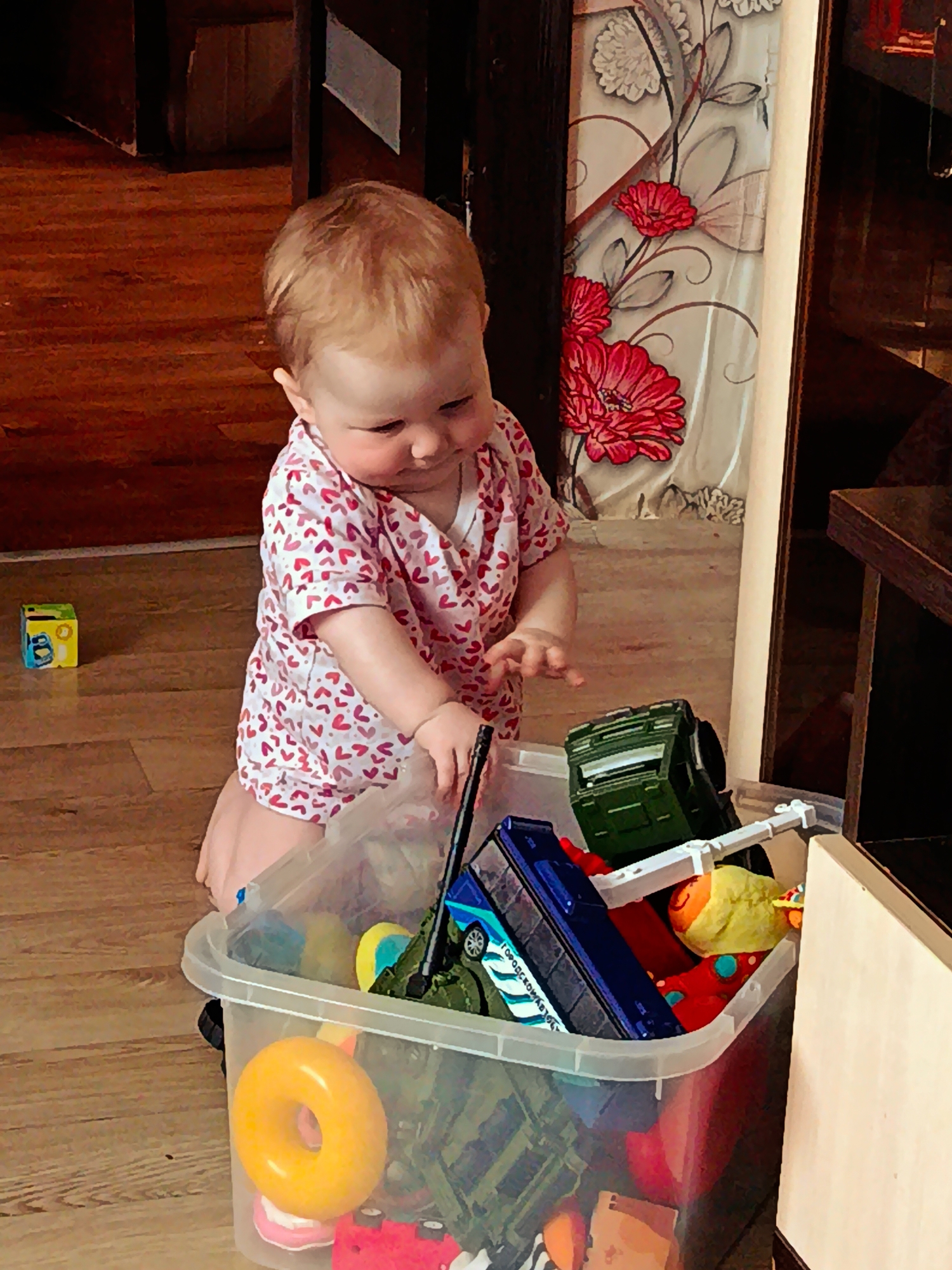 An indoor, eye-level shot captures a baby, likely a toddler, kneeling on a polished wooden floor in a home in Фоки, Russia. The baby, with fair hair and wearing a white short-sleeved onesie adorned with a red heart pattern, is looking down with a focused expression, seemingly reaching into a large, clear plastic toy bin.
The bin is filled with a variety of colorful toys. Prominently visible are a blue toy truck or bus with the text "ГОРОДСКОЙ АВТОБУС" (City Bus) printed on its side, a dark green military-style truck, a bright yellow donut-shaped toy, a red toy vehicle, and an orange fish-like toy. A small yellow building block or cube is seen resting on the wooden floor to the left of the baby.
In the background, a decorative curtain or tapestry featuring large red and grey/white floral and leaf patterns hangs. To the right, dark wooden furniture is partially visible, suggesting a furnished room. The wooden floor exhibits a distinct grain pattern, reflecting warm, bright lighting, which indicates it is daytime. The scene depicts a child engaged in playtime, exploring the contents of their toy bin.