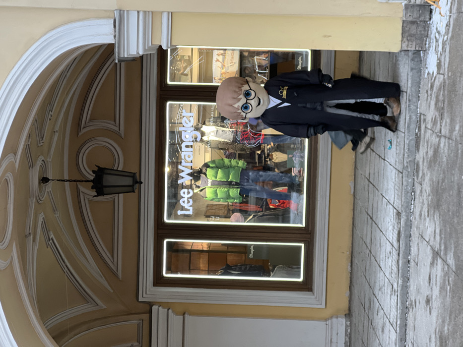 The image depicts an urban outdoor scene in front of a building with a light yellow stucco facade and white architectural trim. An ornate, dark-colored lantern hangs from the decorative ceiling of a prominent arched entryway. Beneath this arch, a retail storefront for "Lee Wrangler" is visible through large glass windows, displaying several mannequins dressed in winter attire, including a bright green puffer jacket and blue jeans.
On the paved sidewalk in the foreground, which is partially covered with patches of dirty snow and ice, stands a person in a full-body mascot costume. The costume is designed to resemble the character Harry Potter, featuring an oversized head with light brown hair, round black-rimmed glasses, and a smiling expression. The body is clad in a black robe with a gold-colored emblem on the left chest area. The wearer's actual brown shoes and lower legs, covered in light-colored pants, are visible beneath the hem of the robe. Black-gloved hands are visible at the end of the robe's sleeves.
Behind the costumed figure, the lower legs and feet of at least two other individuals are partially visible on the sidewalk. A small, bright blue plastic object lies on the ground near the mascot's feet. The architectural style, particularly the ornate arched facade, is characteristic of historic buildings in Saint Petersburg, Russia, and is commonly found along major thoroughfares such as Nevsky Prospect.