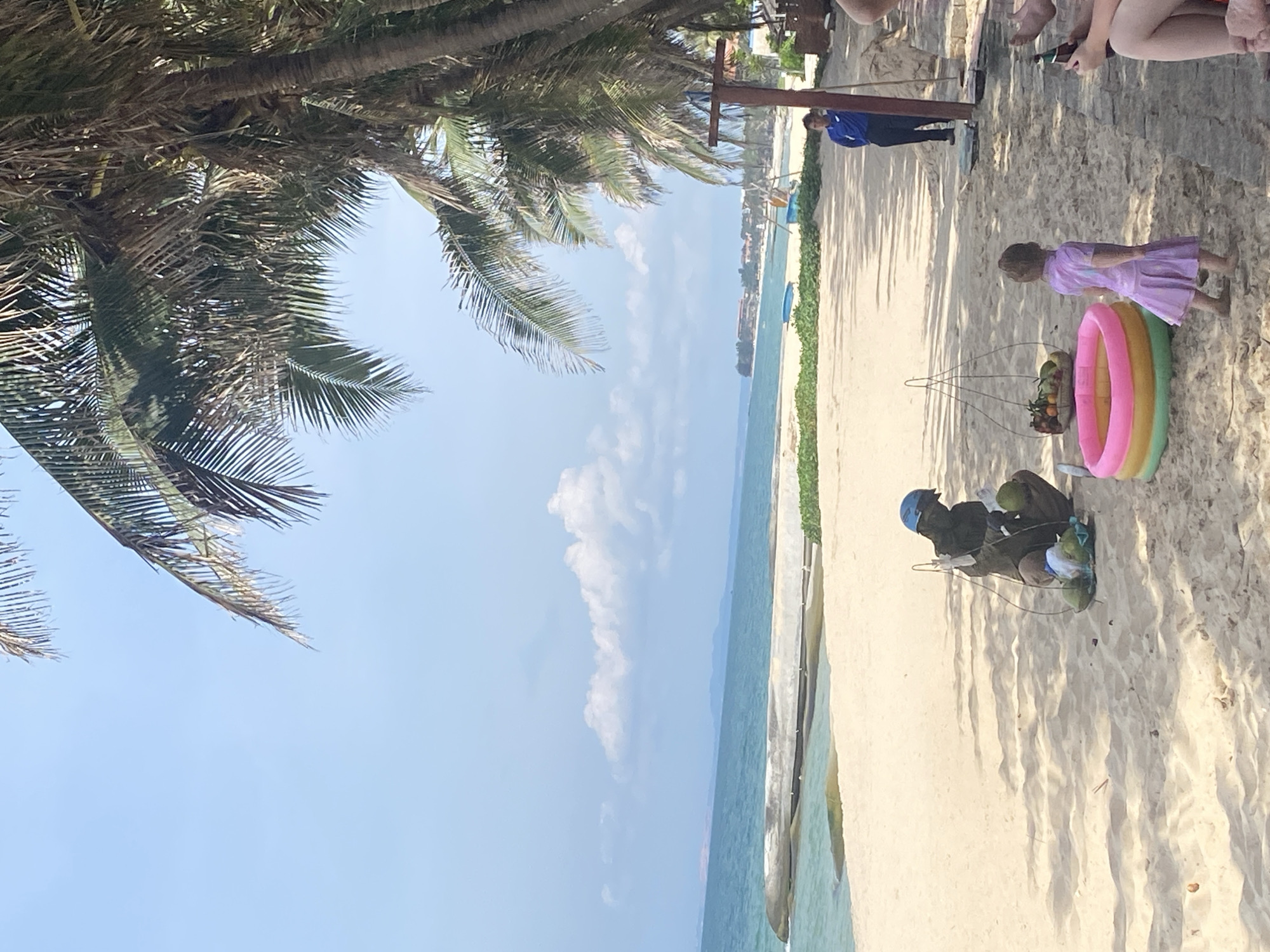 The image depicts a sunny day at a beach in Phu Thuy Ward, Vietnam. Palm trees with fronds hang over the scene, casting shadows on the sand. In the foreground, a person is crouched down preparing coconuts, with a basket of coconuts at their side. Next to them is a small inflatable pool filled with water and a basket containing fruits. A young girl in a purple dress is standing near the pool, facing away from the camera. Further in the background, a person in a blue uniform stands near a structure. The ocean with gentle waves is visible beyond the sandy beach, and the sky is a clear blue with scattered white clouds. Buildings and vegetation can be seen in the distance along the coastline.