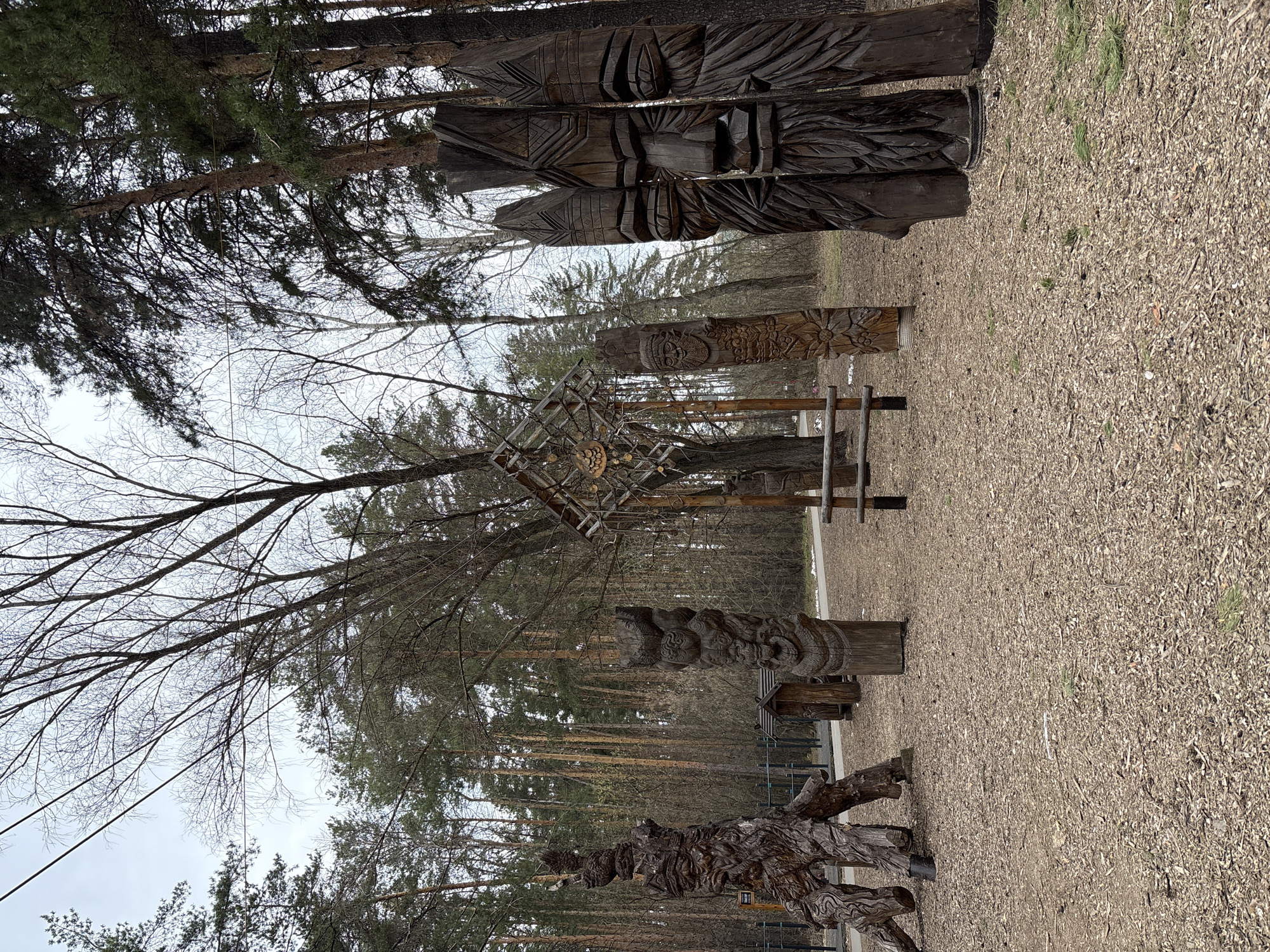The image depicts a collection of wooden carvings situated in a park or wooded area, likely in Neftekamsk, Russia. Tall, carved wooden poles with faces and figures stand prominently. Other carved wooden structures, including a geometric, box-like design and a stylized animal or mythical creature, are also present. The ground is covered in wood chips or mulch. Trees surround the carvings, creating a natural backdrop. The sky is overcast. No people are visible in the frame.