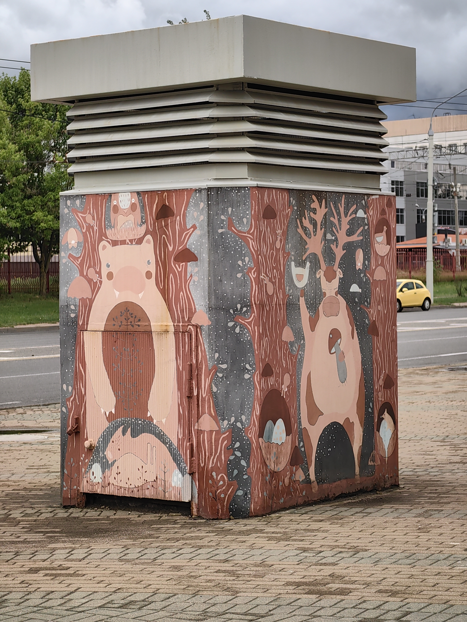 This media file shows a rectangular, gray metal utility box, likely an electrical substation, adorned with vibrant, whimsical murals. The artwork depicts fantastical forest creatures and elements in a soft, pastel color palette of pinks, browns, and grays.
On the left side of the box, a large, pink bear-like creature with tusks is centrally positioned, framed by stylized tree trunks and scattered mushrooms. Above it, a small owl-like creature sits in a nest. Below the bear, a sleeping rabbit is curled up under a rainbow.
The right side of the box features a large, pink deer-like creature with impressive antlers, also surrounded by tree motifs. This creature holds a small bird in one of its hands and appears to be interacting with a mushroom. Other smaller creatures and mushroom shapes are scattered throughout the mural.
The utility box is situated on a paved walkway made of rectangular interlocking stones, which appear slightly damp, suggesting recent rain or high humidity. In the background, a road is visible with a yellow car driving by. Beyond the road, there are buildings, some with windows and scaffolding, and trees. Power lines and a lamppost are also visible, indicating an urban or suburban setting. The sky is overcast with thick, gray clouds, suggesting a cloudy or possibly rainy day. There is no visible text in the image. The overall impression is one of colorful, playful street art transforming a utilitarian object into a point of interest in an everyday urban landscape.