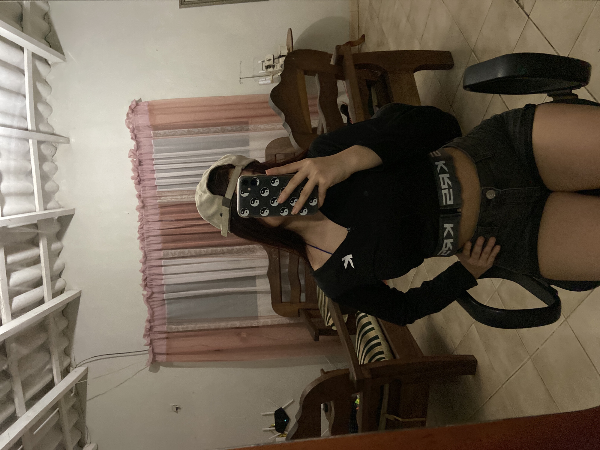 A person is sitting on a rolling office chair indoors, facing away from the camera. They are taking a picture of themselves in a mirror with their phone. The person is wearing a beige cap, a black long-sleeved shirt, and dark denim shorts with a black belt. The phone case has a yin-yang symbol pattern. The background includes wooden furniture (a chair and a small table) and a window with blinds. The floor appears to be tiled. While a specific location in Juan Griego, Venezuela cannot be determined from the image, it appears to be a residential interior.