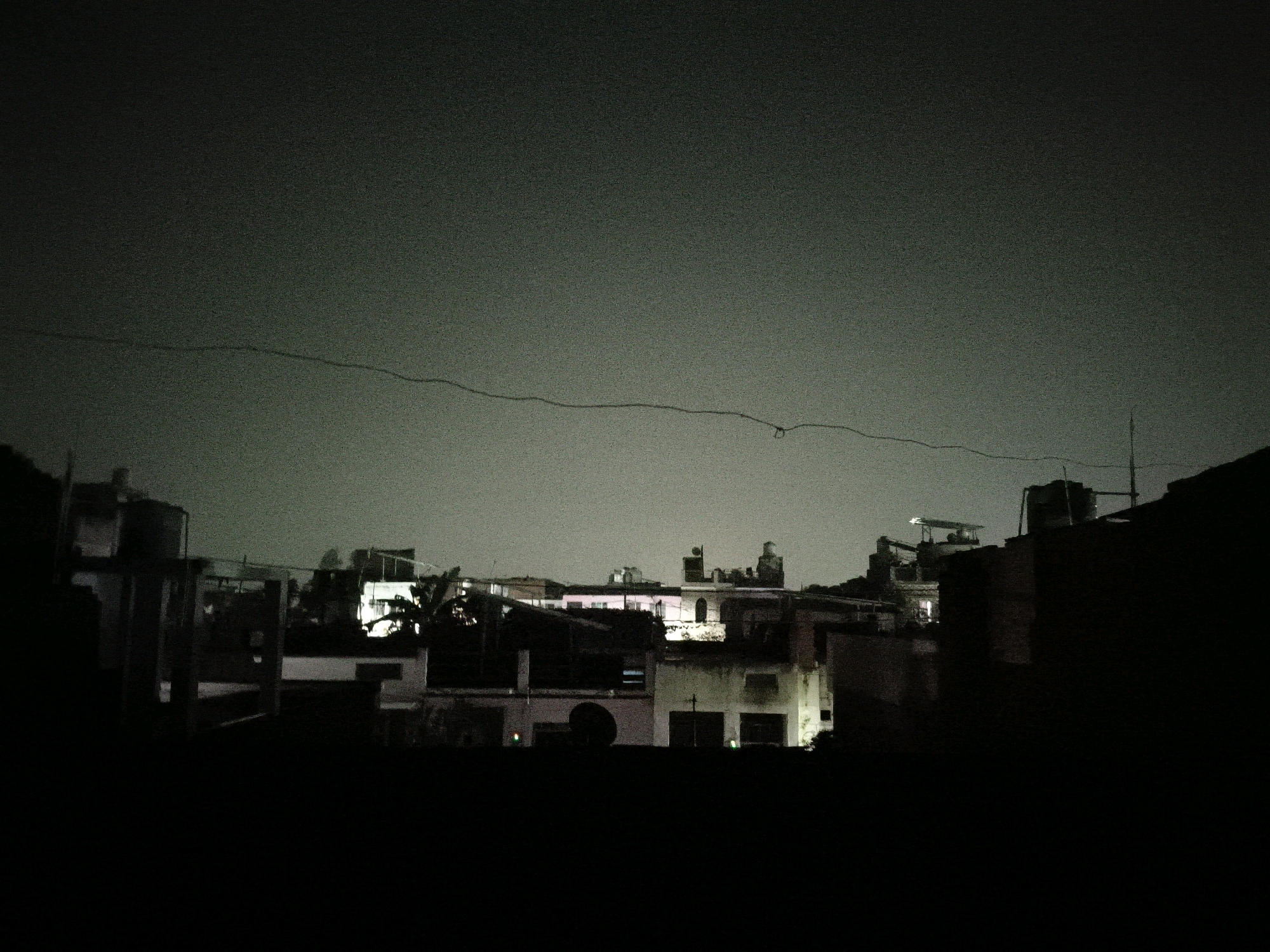 The image depicts a nighttime urban scene, likely from a rooftop in the city of Bareilly, India. The sky is dark and overcast, with a faint, grainy texture indicating a high ISO setting or low light conditions. A thin, dark wire stretches horizontally across the upper portion of the frame.
Below the sky, a silhouette of buildings and structures is visible, illuminated by some artificial lights. The architecture appears to be a mix of residential and commercial buildings, with some showing signs of wear. Several windows emit light, suggesting occupancy. One prominent building on the left side has a cylindrical water tank on its roof. In the center, a building is partially lit, with some parts appearing white and others darker. There's a hint of pink light emanating from some windows in the middle ground. On the right, a darker, more imposing structure is partially visible, obscuring the background. The foreground is almost entirely black, suggesting the viewpoint is from a dark area, possibly another rooftop or elevated platform. No people are visible in the image. The overall impression is one of a quiet, dimly lit urban environment at night.