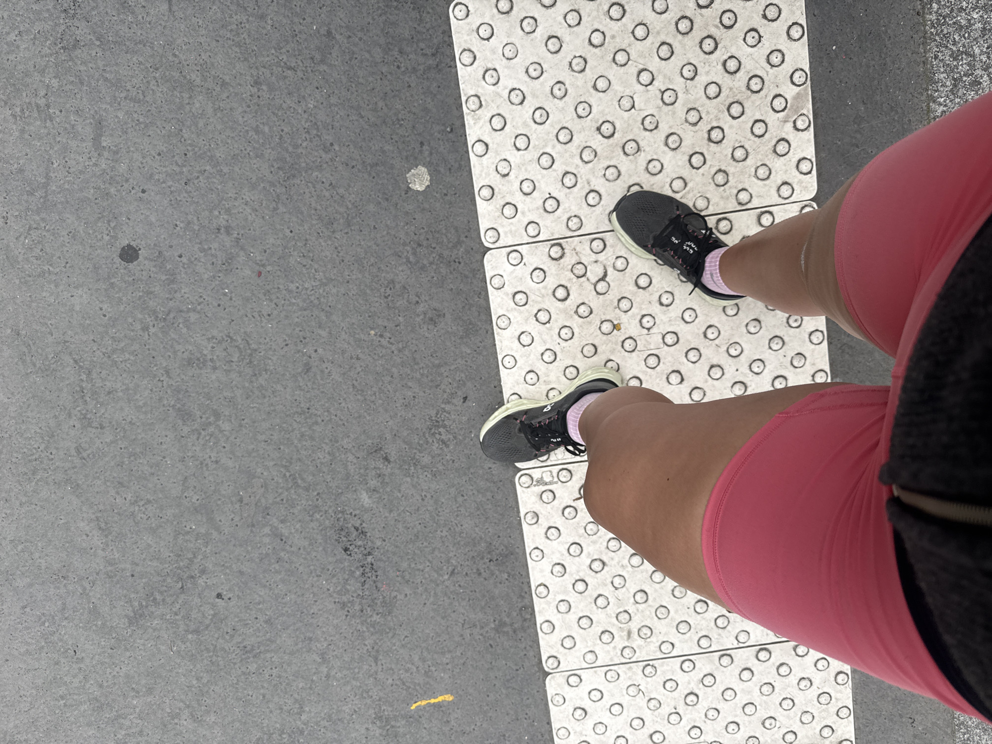 The image shows a view from above, likely taken by a person looking down at their own legs and feet. The person is wearing pink shorts, black running shoes, and pink socks. Their legs are positioned on a section of white tactile paving tiles, designed with raised circular dots for pedestrian guidance. To the left of the tactile paving is a gray concrete surface. The location is likely an urban area in Paris, France, given the prevalence of tactile paving for accessibility in pedestrian zones. There are no other visible people or objects. The focus is on the person's legs and feet in relation to the tactile paving.